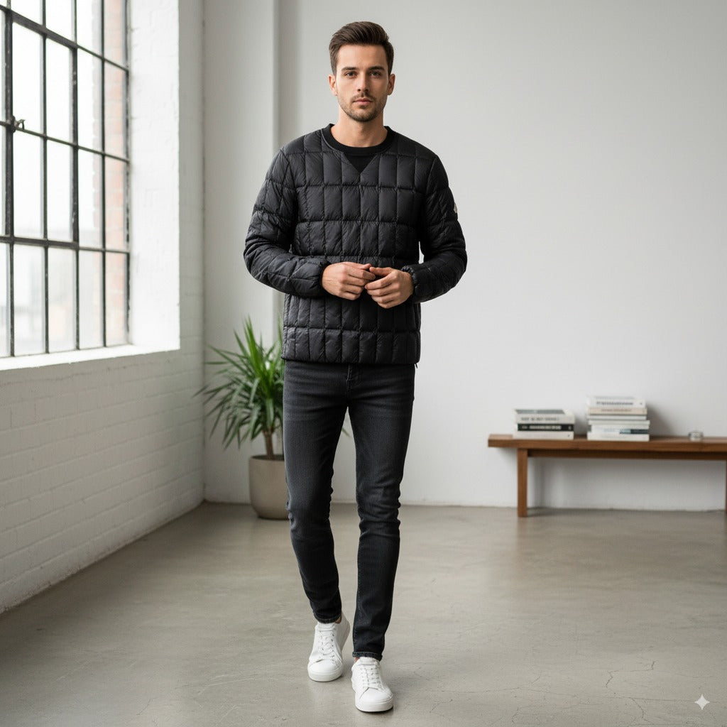 Ein Mann in einer schwarzen Steppjacke, schwarzen Jeans und weißen Turnschuhen steht in einem Innenraum auf einem polierten Betonboden, mit einem großen Fenster, einer Topfpflanze und einer Bank mit gestapelten Büchern im Hintergrund.