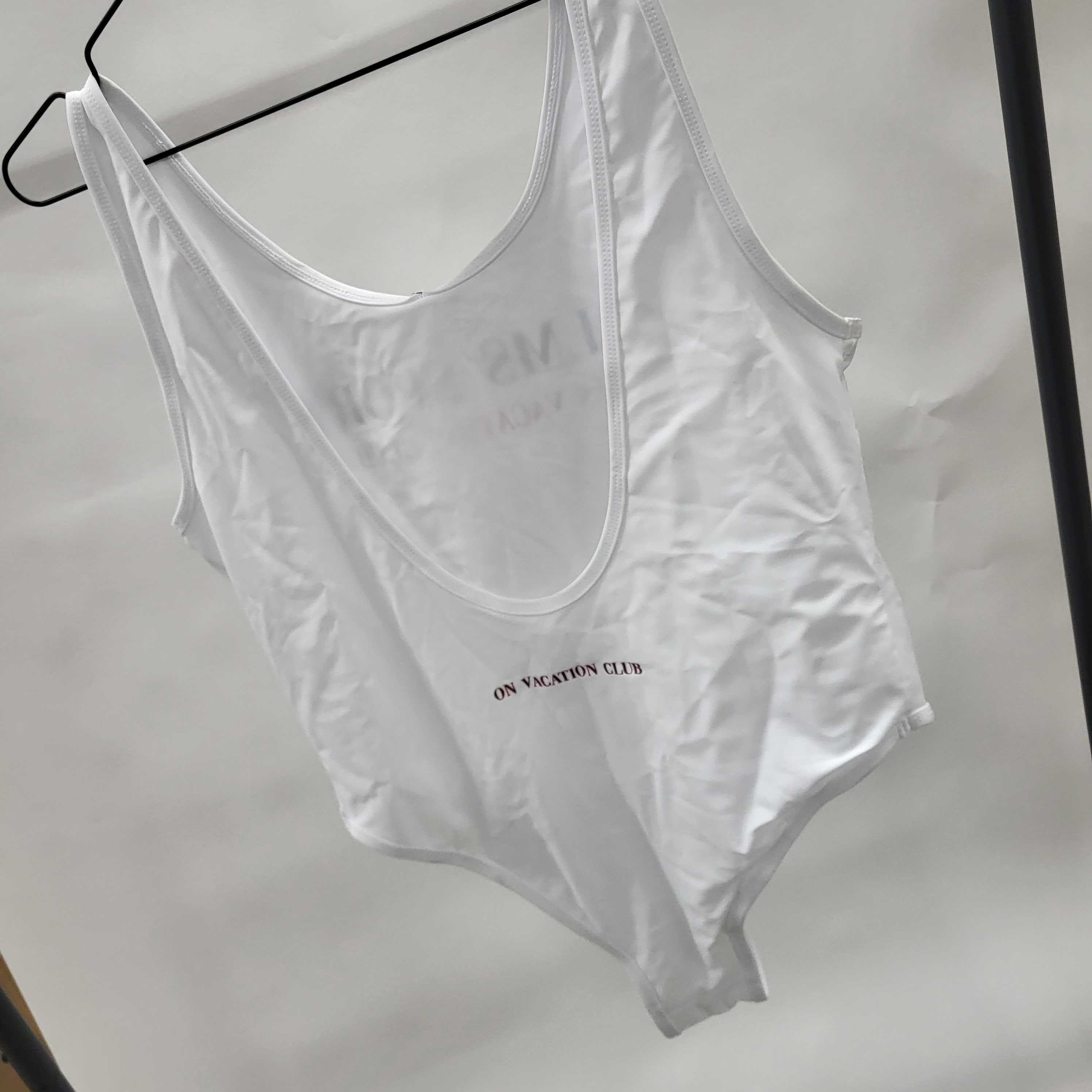 Weißer Badeanzug mit dem Aufdruck "ON VACATION CLUB" hängt auf einem Kleiderbügel vor einem neutralen Hintergrund.