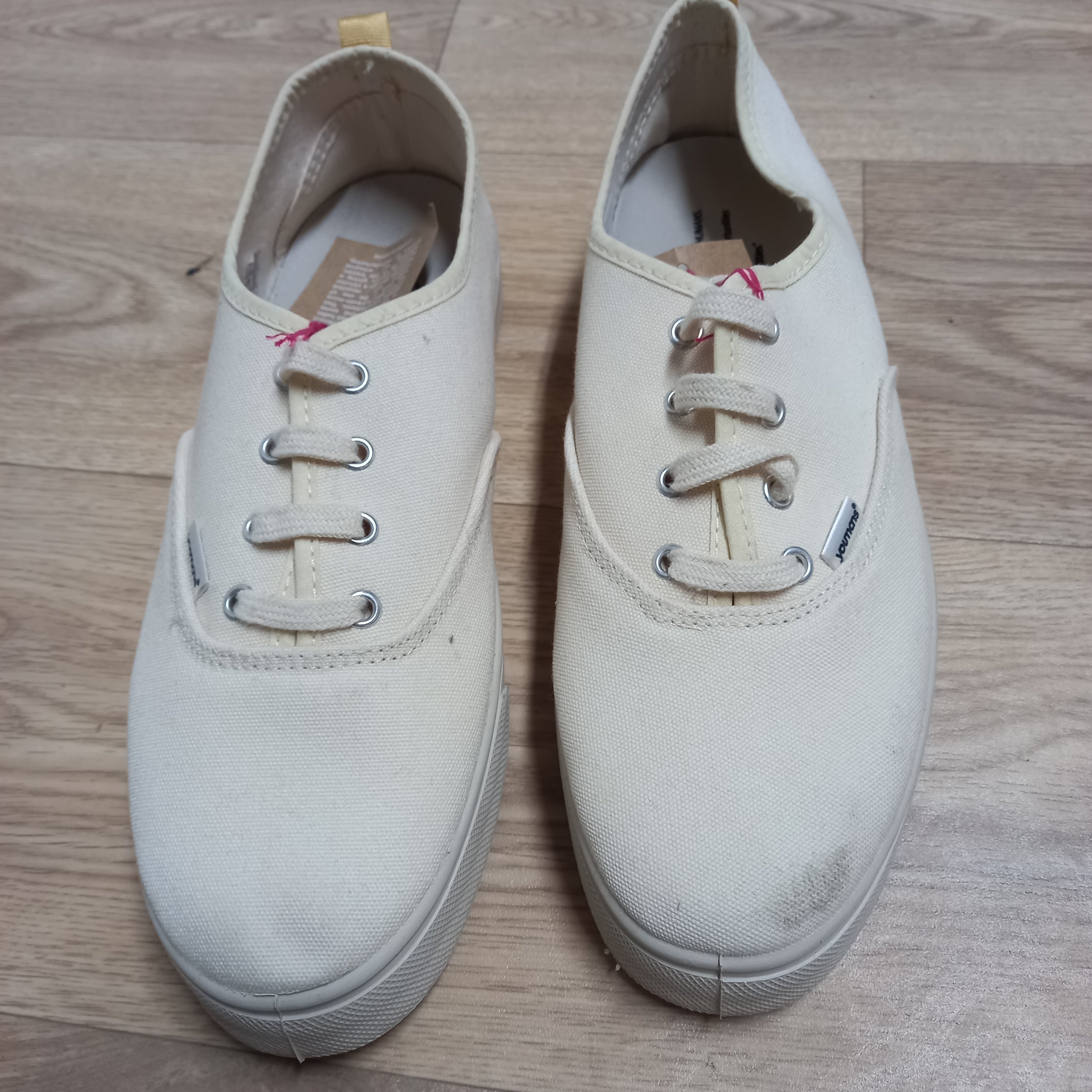 Zwei cremefarbene Turnschuhe mit Schnürsenkeln stehen auf einem hellen Holzboden.