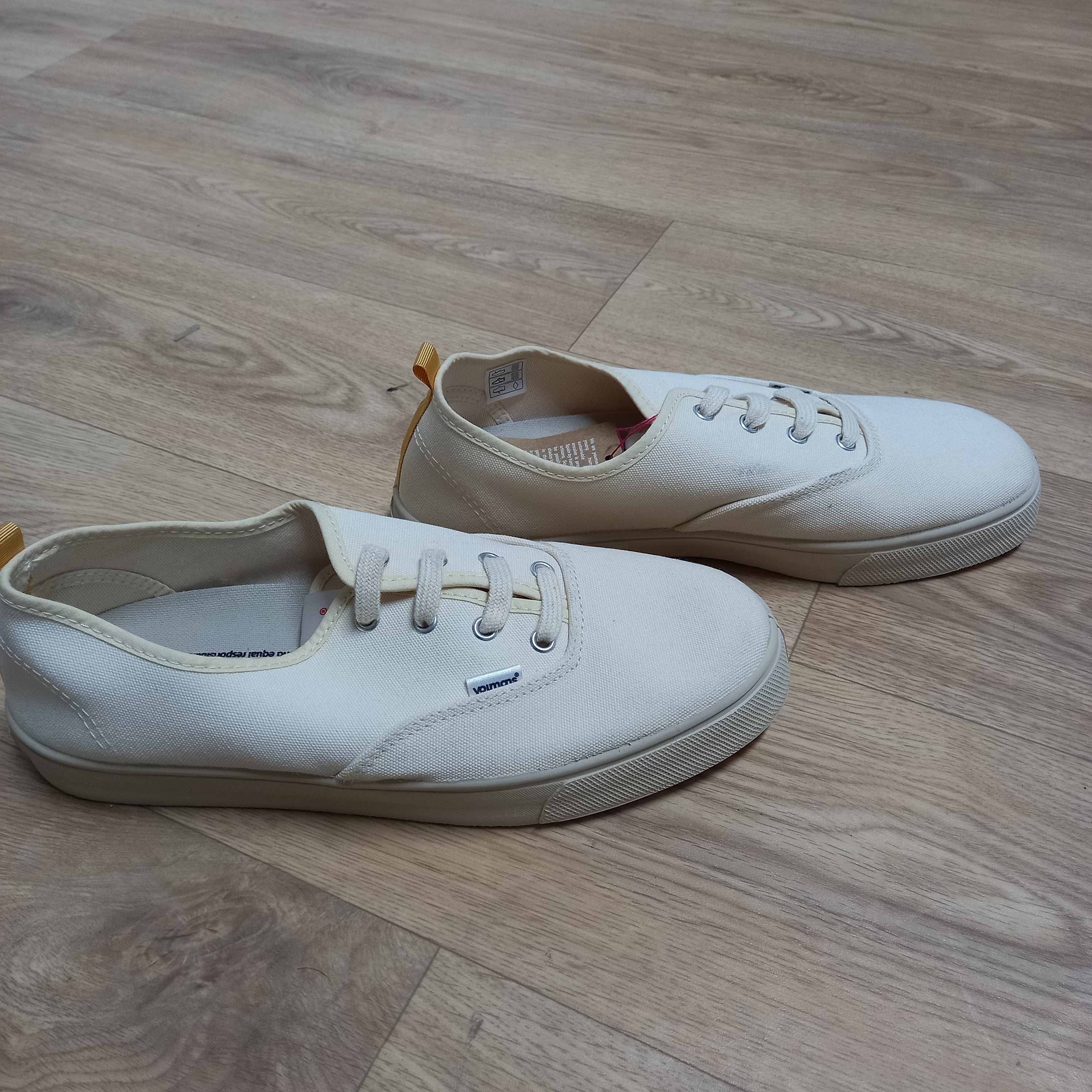 Weiße Segeltuchschuhe mit Schnürsenkeln auf einem hellen Holzboden.