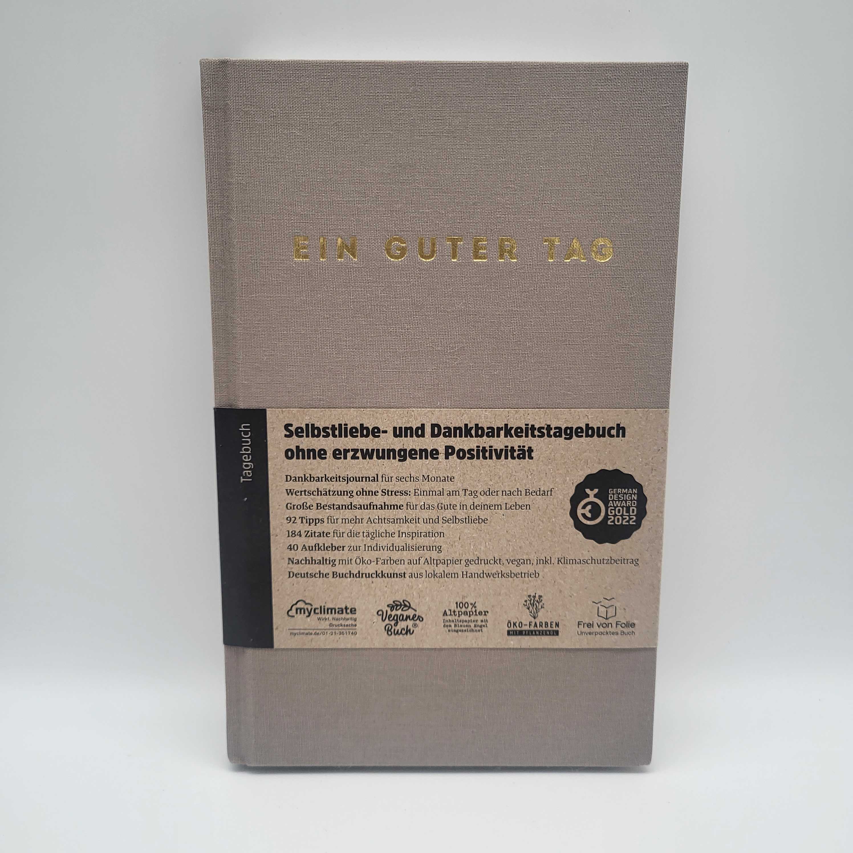 Graues Tagebuch mit der Aufschrift "Ein Guter Tag" und Papierbanderole mit Informationen zur Dankbarkeit und Selbstliebe.