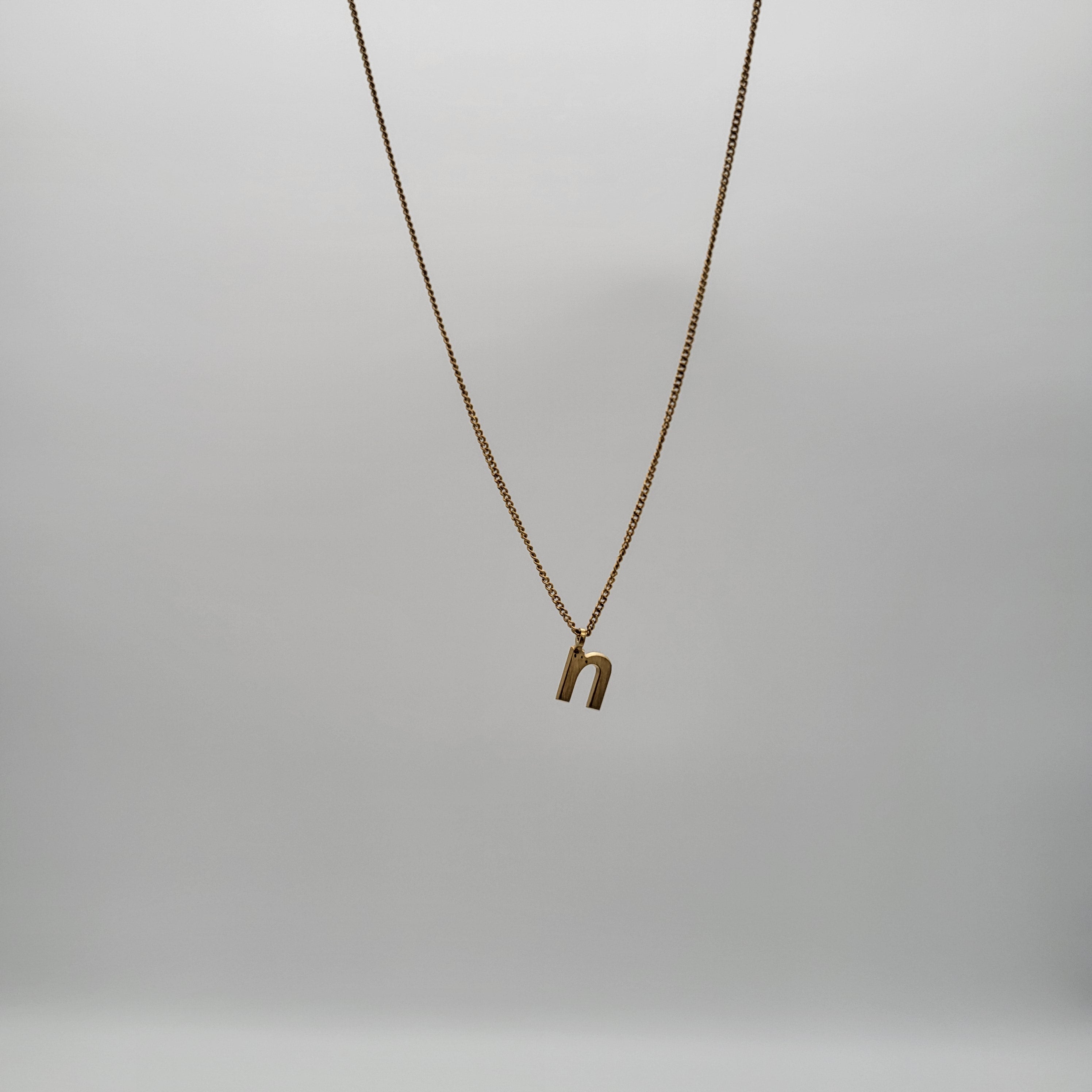 Eine goldene Halskette mit einem Anhänger in Form des Kleinbuchstabens n hängt vor einem schlichten, hellgrauen Hintergrund.