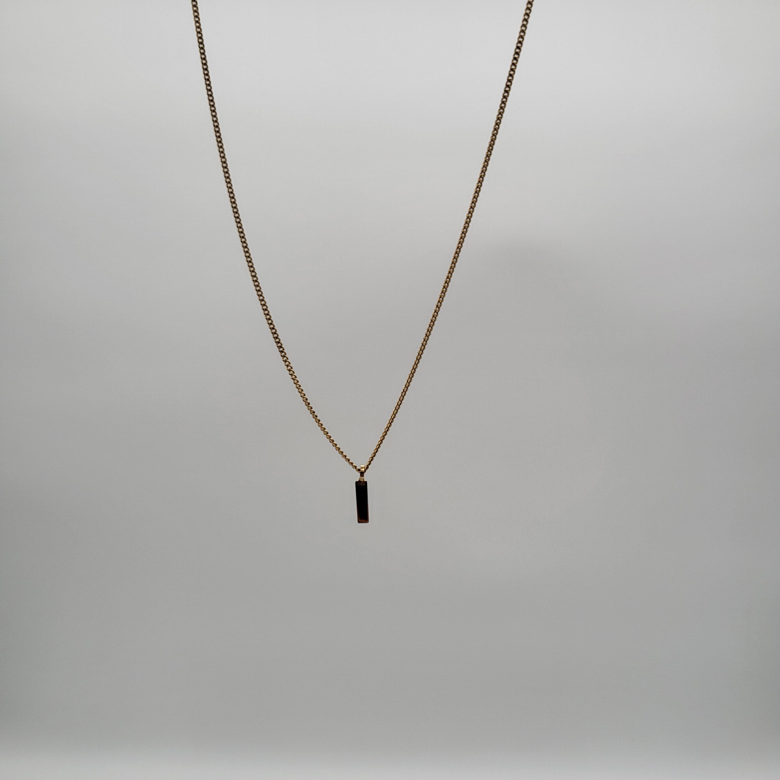 Eine goldene Halskette mit einem kleinen, rechteckigen schwarzen Anhänger hängt vor einem schlichten, hellgrauen Hintergrund.