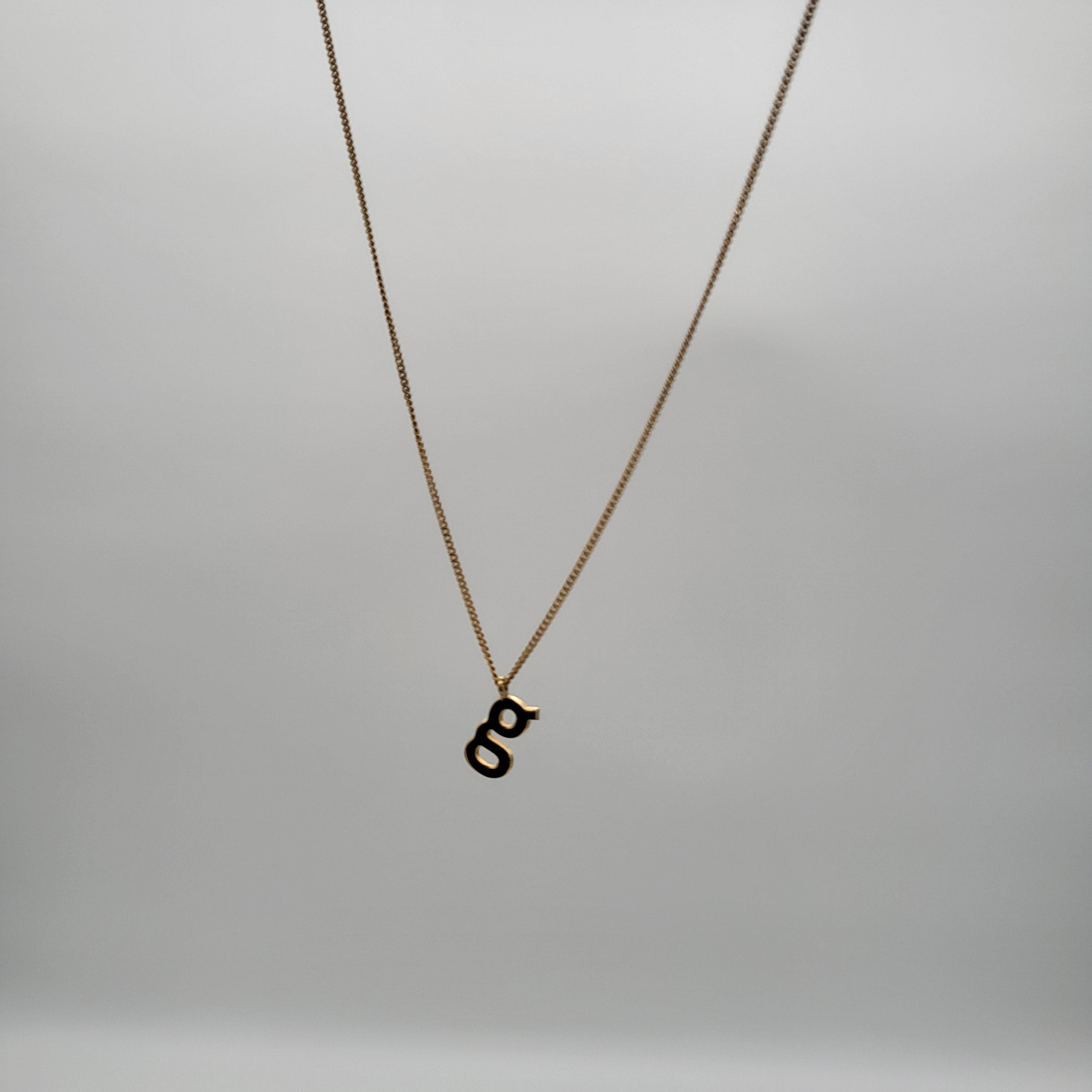 Eine zarte Goldkette mit einem Kleinbuchstaben g als Anhänger vor einem schlichten hellgrauen Hintergrund.