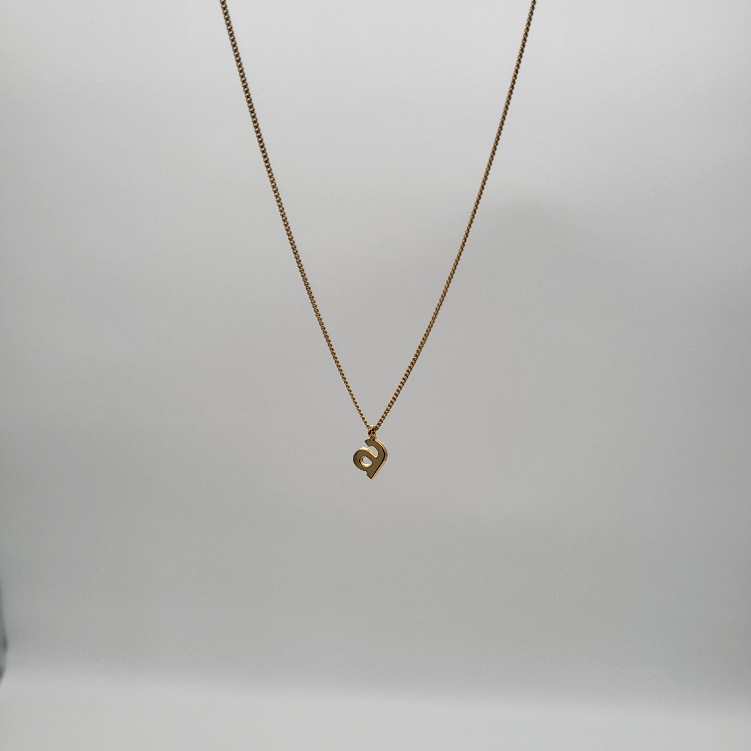 Eine goldene Halskette mit einer zarten Kette und einem kleinen Anhänger in Form des Buchstabens a, der vor einem schlichten weißen Hintergrund hängt.