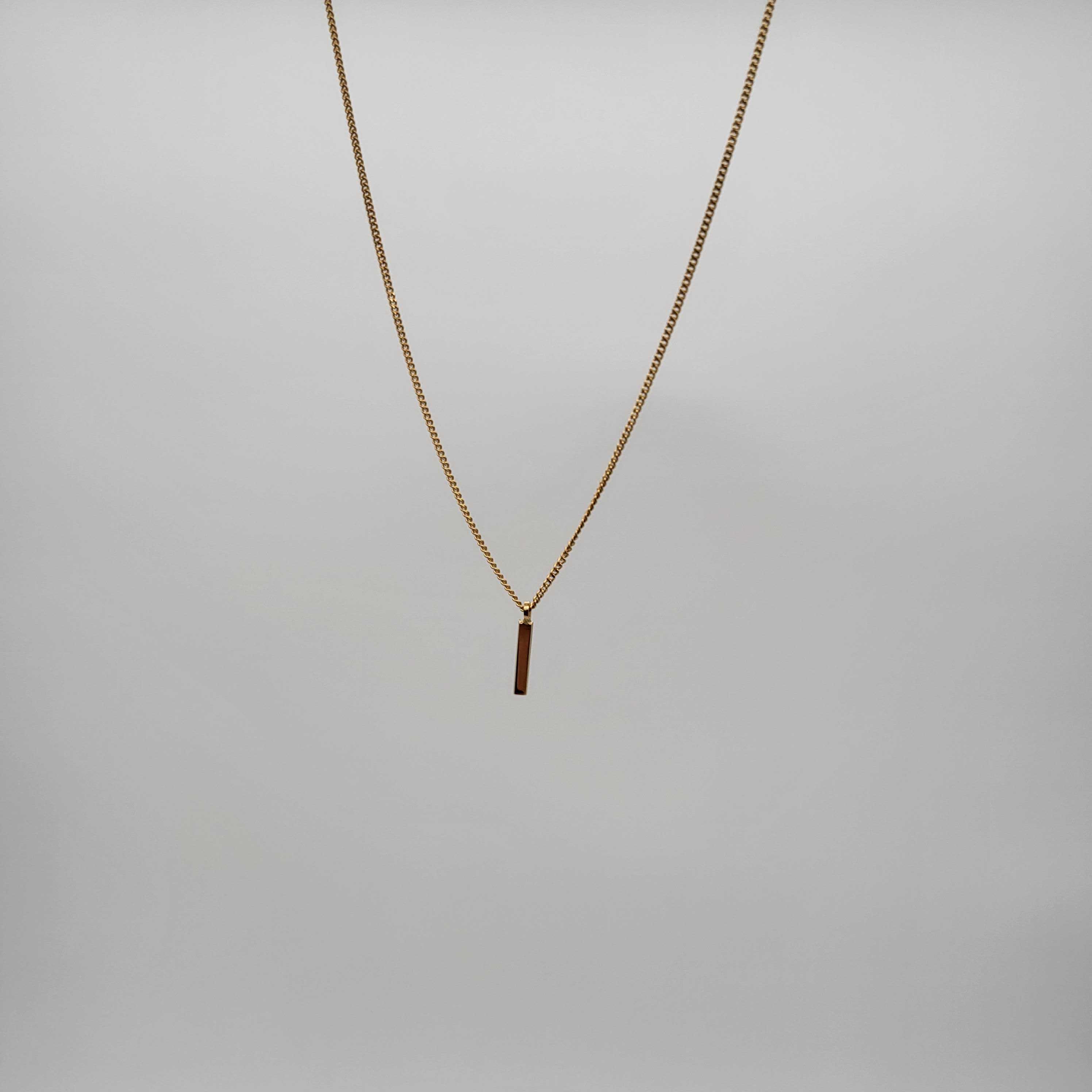 Eine zarte Goldkette mit einem dünnen, vertikalen, rechteckigen Anhänger in der Mitte, der vor einem schlichten hellgrauen Hintergrund steht.