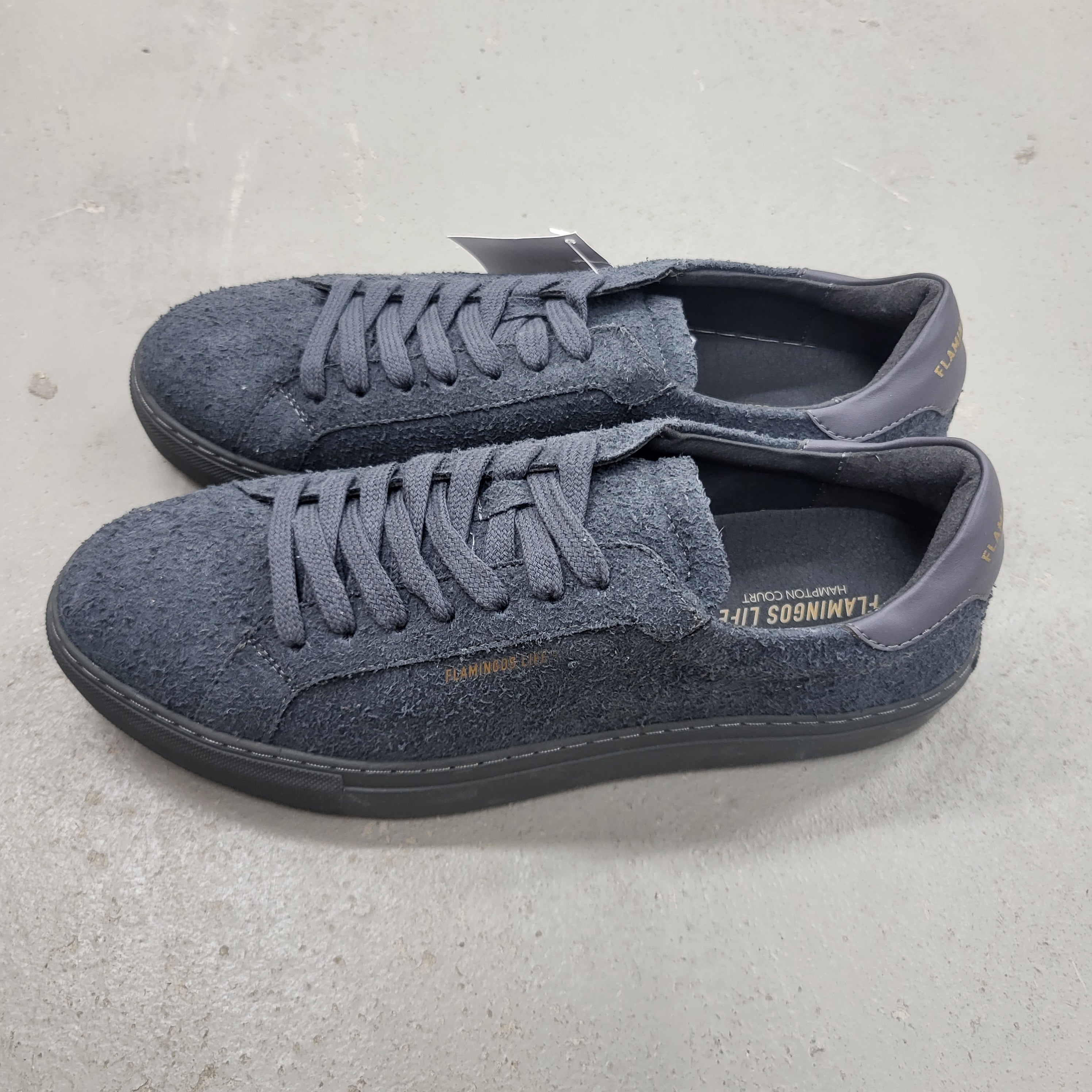 Ein Paar dunkelgraue Wildleder-Sneaker mit passenden Schnürsenkeln und schwarzen Sohlen, die nebeneinander auf einem hellgrauen Betonboden stehen. Die Innenseite und die Fersenlasche haben einen goldenen Branding-Text.