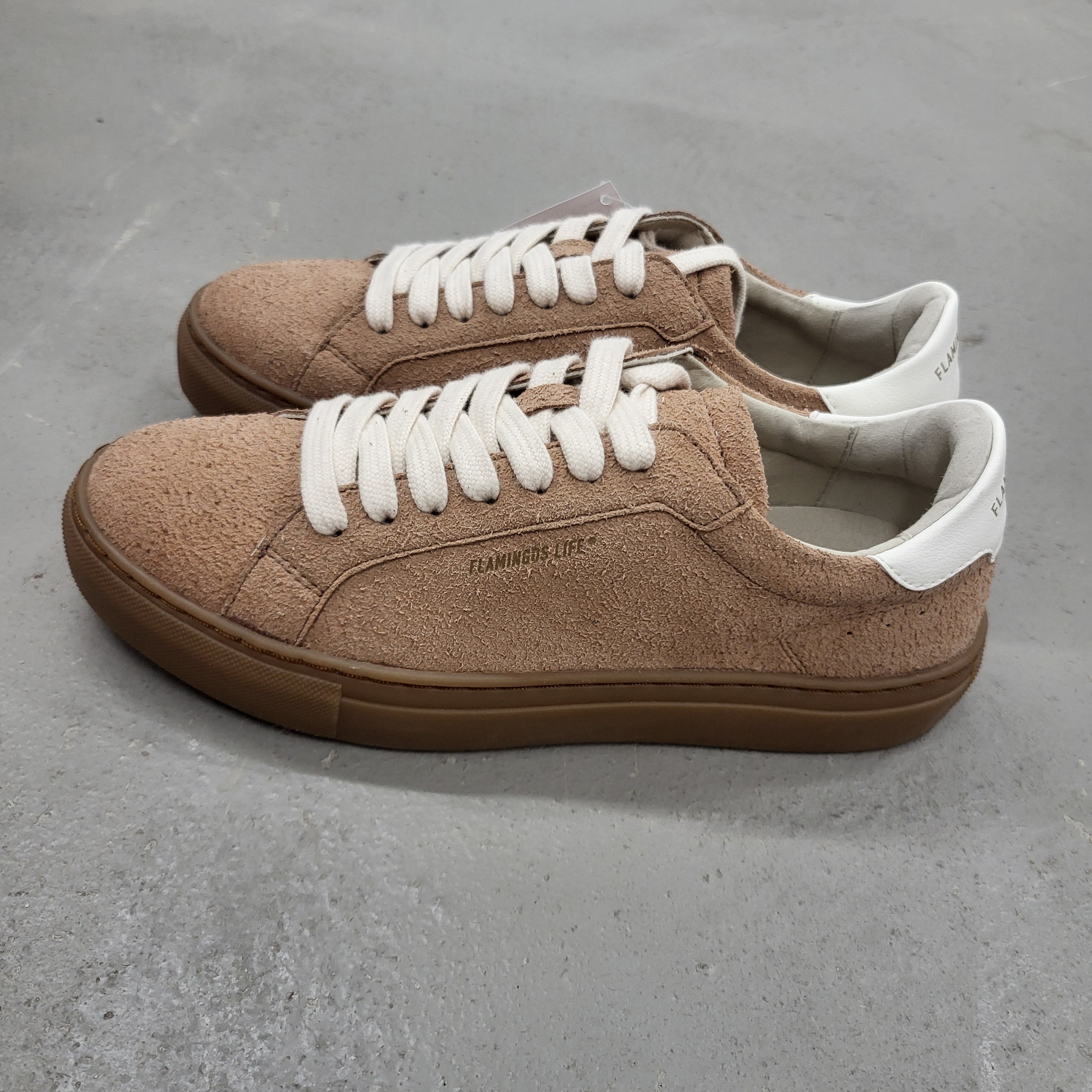 Ein Paar hellbraune Wildleder-Sneakers mit weißen Schnürsenkeln und weißen Fersenlaschen, die auf einer grauen Betonfläche stehen. Die Schuhe haben braune Gummisohlen und ein dezentes Branding an der Seite.