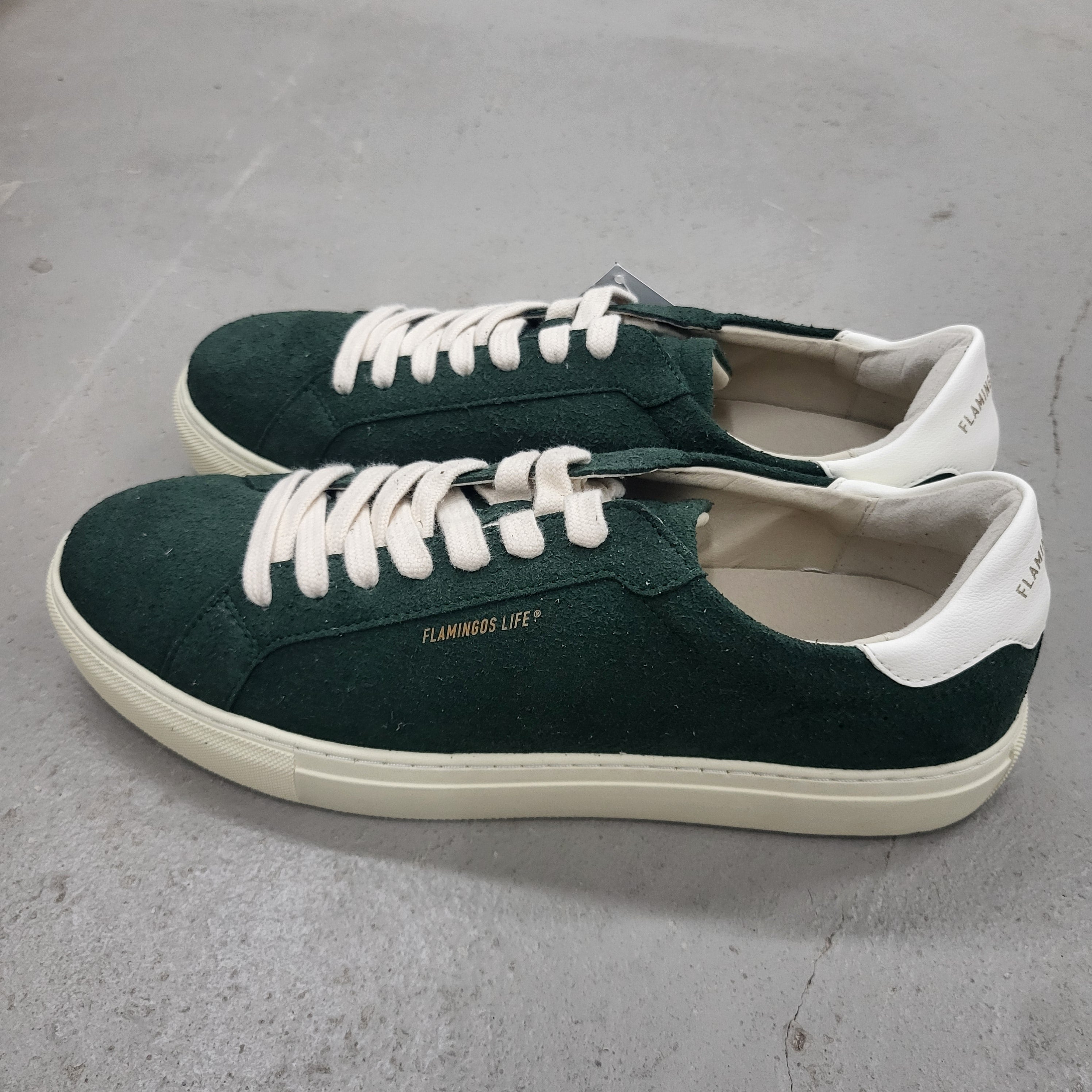 Ein Paar dunkelgrüne Wildleder-Sneakers mit cremefarbenen Sohlen, weißen Schnürsenkeln und weißen Fersenkappen. Auf der Seite der Schuhe steht in goldenen Lettern FLAMINGOS LIFE. Die Schuhe stehen auf einem grauen Boden.