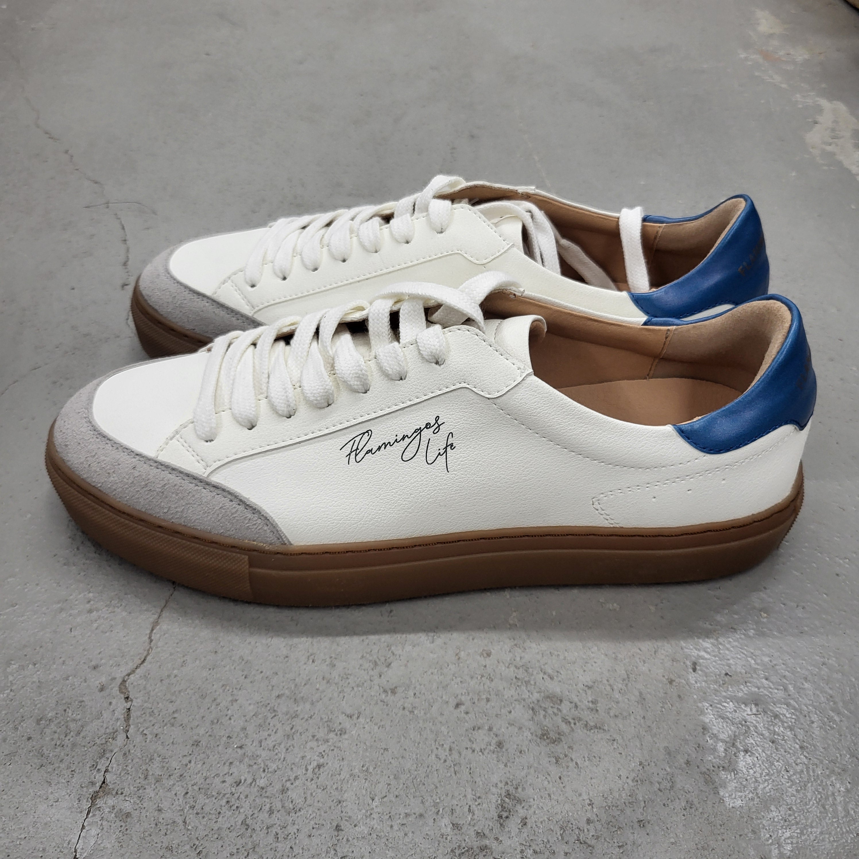 Ein Paar weiße Low-Top-Sneaker mit grauen Zehenkappen, blauen Absätzen, braunen Gummisohlen und der Aufschrift "Running Life" in Schreibschrift an der Seite. Die Schuhe stehen auf einem grauen Betonboden.