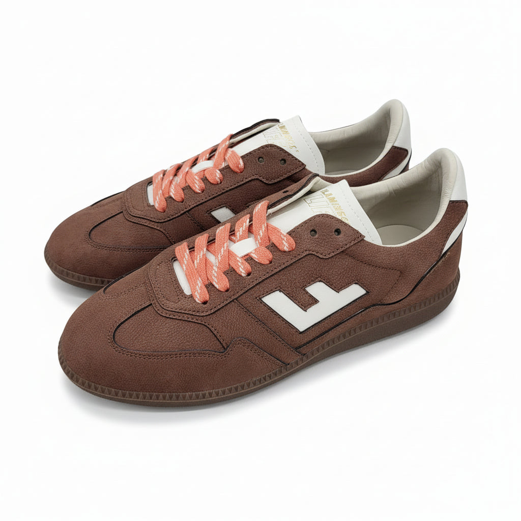 Ein Paar braune Wildleder-Sneakers mit weißen Akzenten, auffälligen weißen F-Logos an den Seiten und leuchtend orangefarbenen Schnürsenkeln, die auf einer grauen Betonfläche stehen.