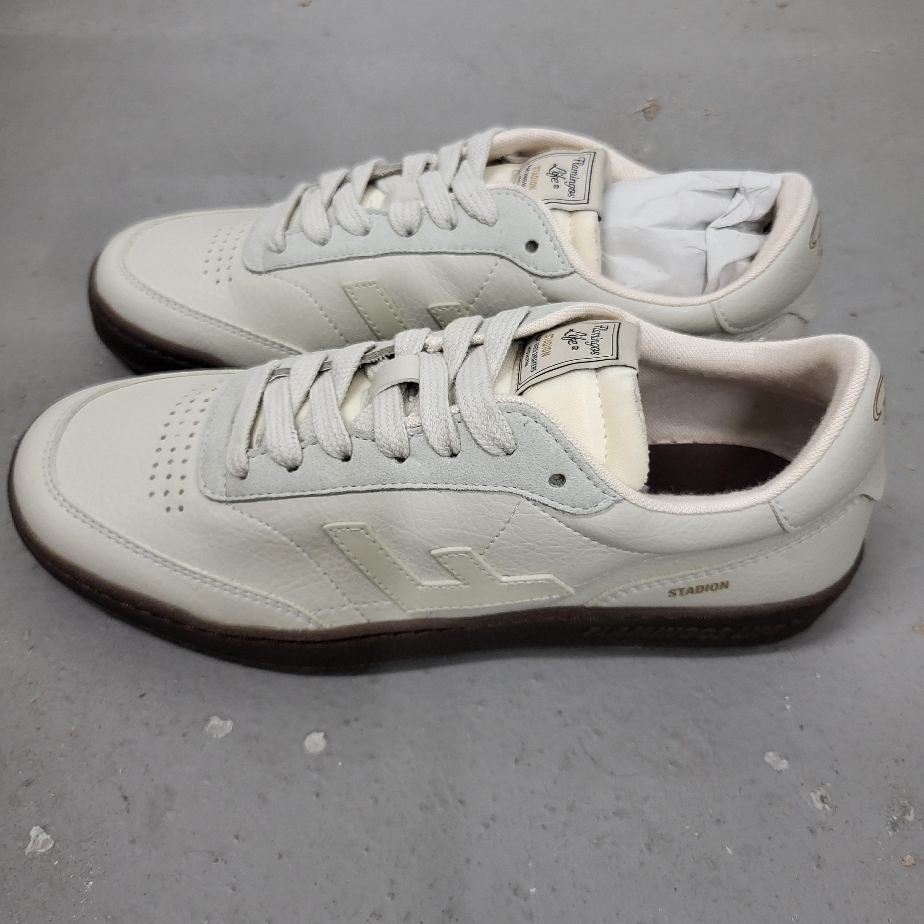 Ein Paar weiße Lederschuhe mit beigen Schnürsenkeln und braunen Sohlen stehen nebeneinander auf einer grauen Fläche. Die Schuhe haben ein stilisiertes L-Logo und das Wort STADION an der Seite.