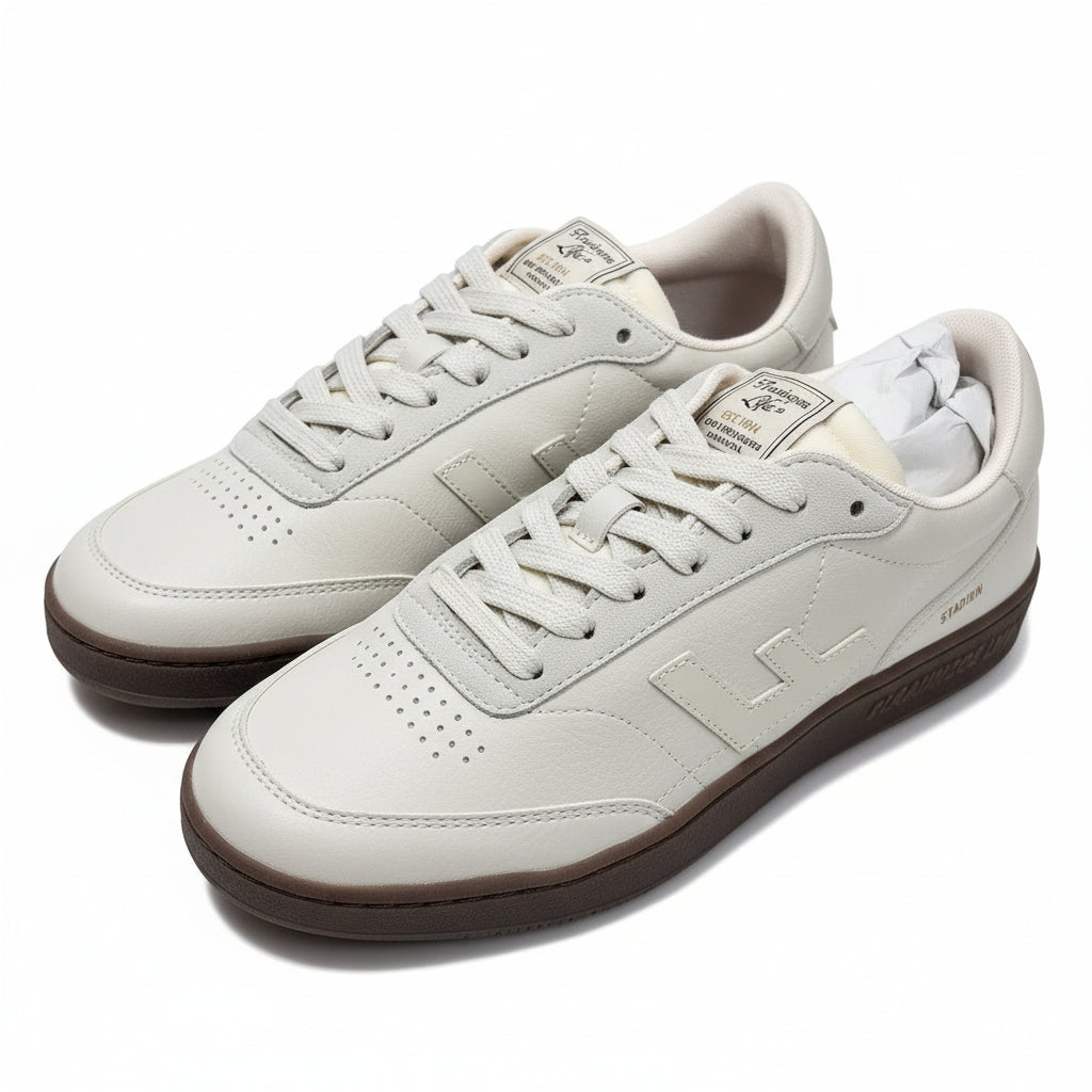 Ein Paar weiße Lederschuhe mit beigen Schnürsenkeln und braunen Sohlen stehen nebeneinander auf einer grauen Fläche. Die Schuhe haben ein stilisiertes L-Logo und das Wort STADION an der Seite.