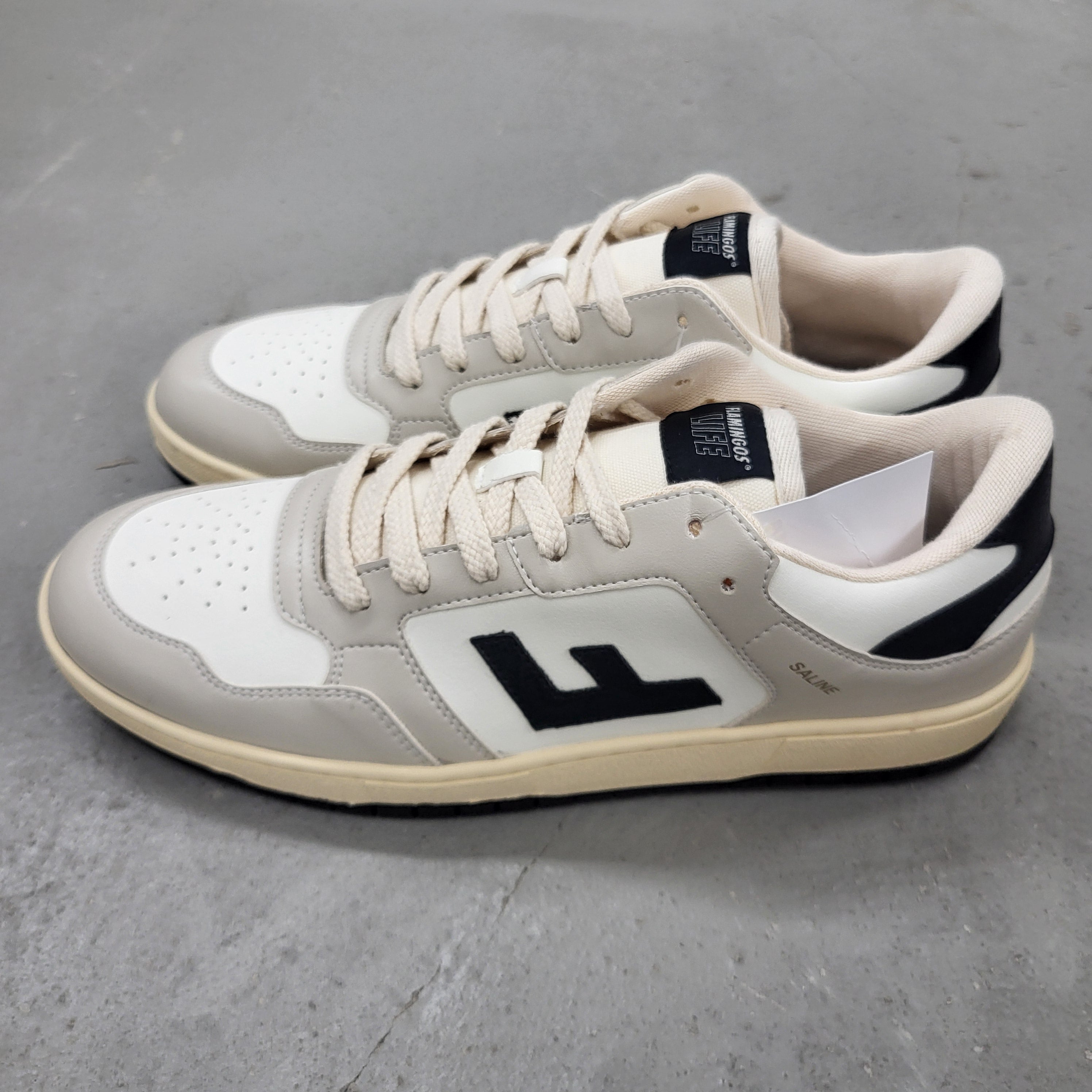 Ein Paar Turnschuhe mit cremefarbenen Schnürsenkeln, weißem Obermaterial, hellgrauen Akzenten, schwarzer Laufsohle und einem großen schwarzen F-Logo an der Seite, die auf einem grauen Betonboden stehen.