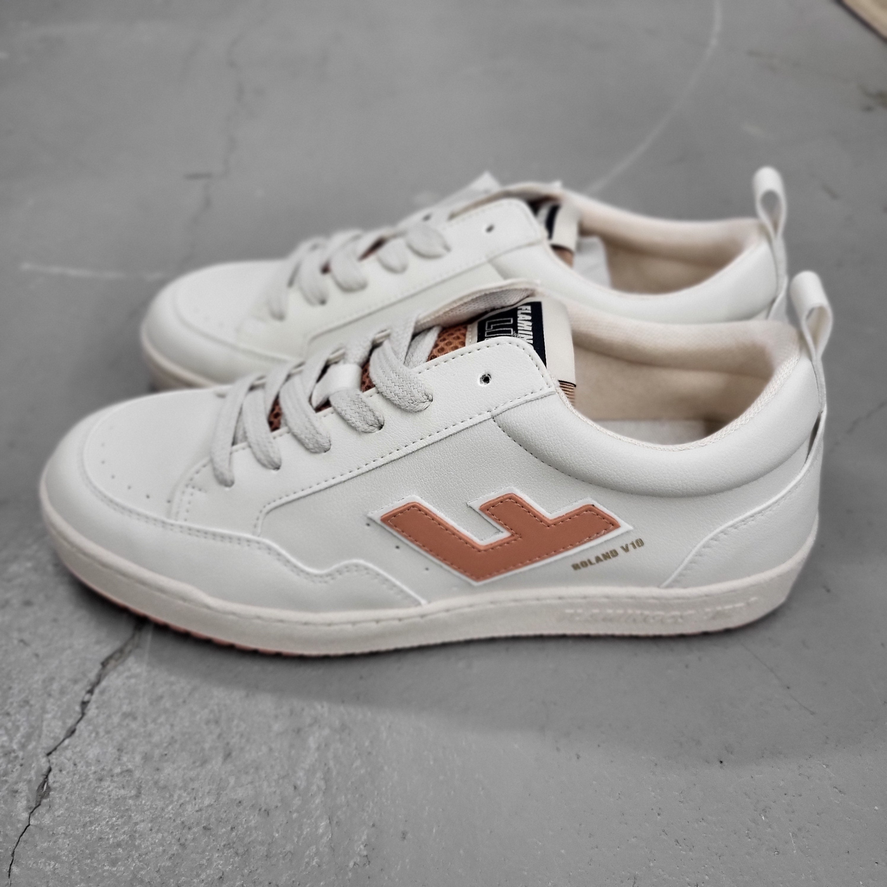 Ein Paar weiße Turnschuhe mit hellbrauner Sohle und einem großen hellbraunen L-Logo an der Seite, die auf einem grauen, leicht rissigen Betonboden stehen.