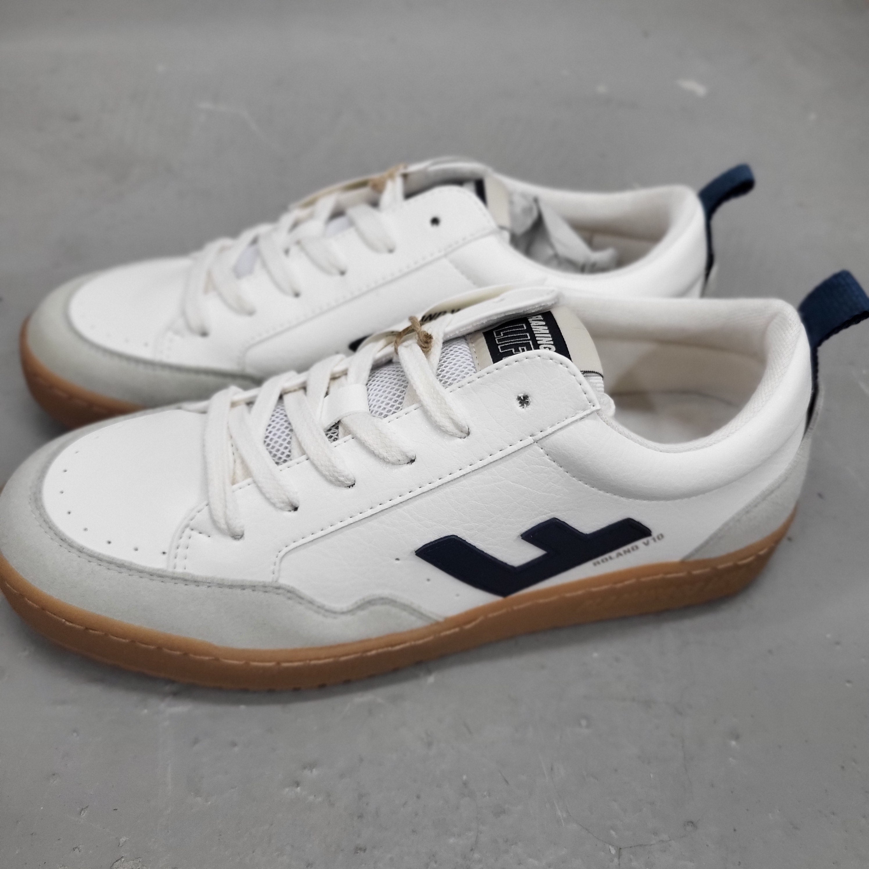 Ein Paar weiße und graue Turnschuhe mit Gummisohlen, marineblauen Fersenlaschen und einem großen marineblauen L-Logo an der Seite, die auf einem grauen Boden stehen.