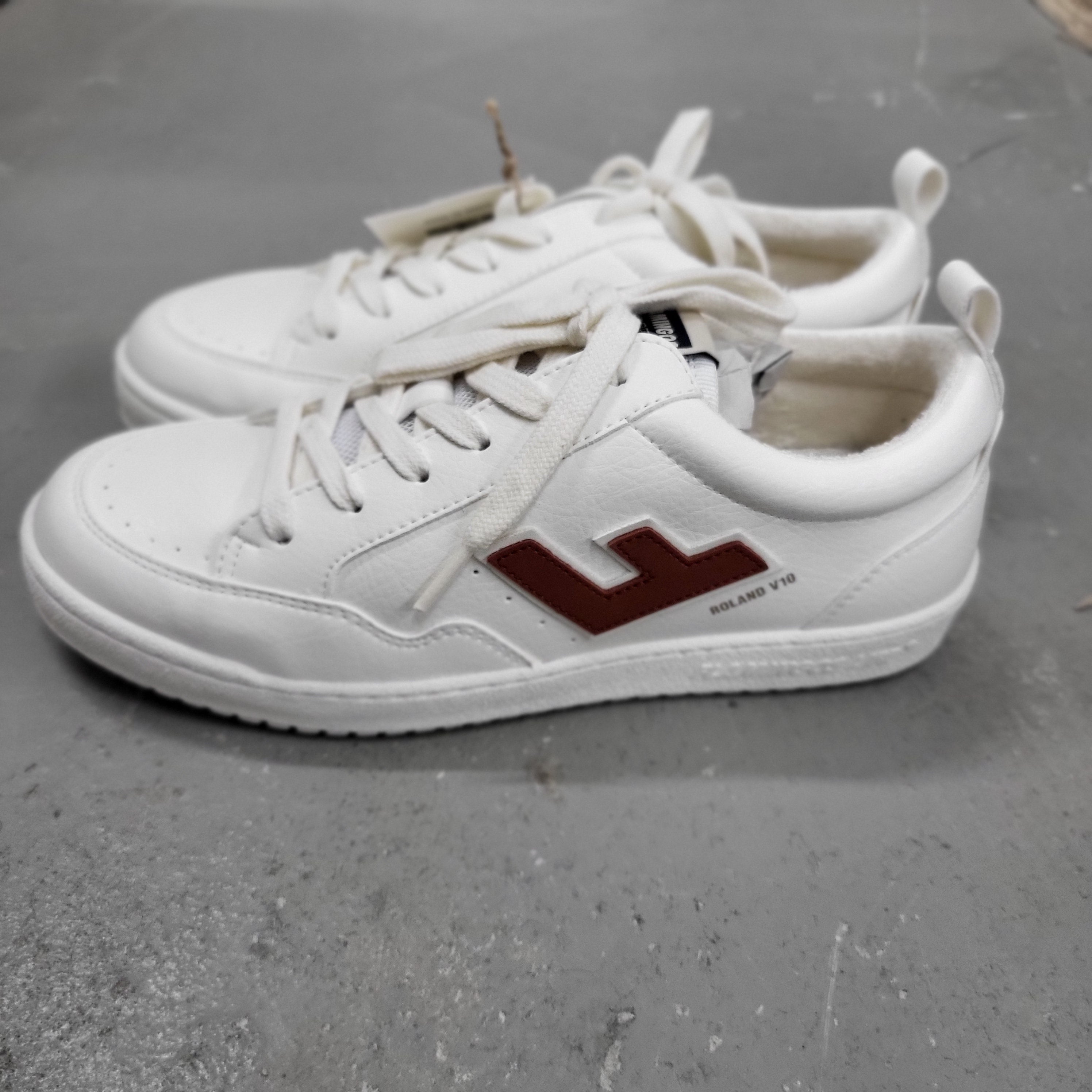 Ein Paar weißer Turnschuhe mit braunen seitlichen Logo-Akzenten und weißen Schnürsenkeln, die auf einer grauen Betonfläche stehen.