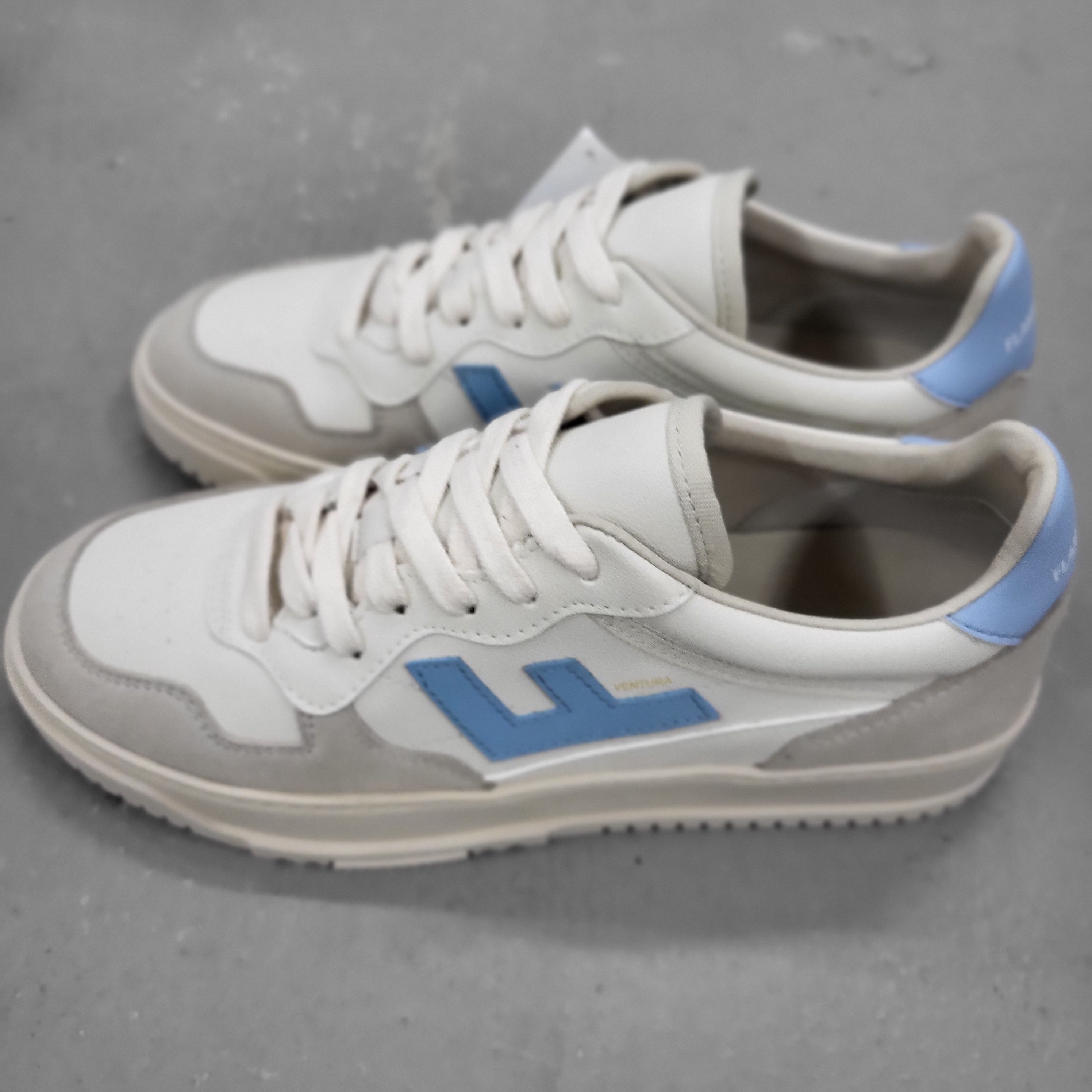 Ein Paar weiße und hellgraue Turnschuhe mit hellblauen Akzenten, einschließlich eines seitlichen Logos und einer Fersenlasche, auf einer grauen Oberfläche.