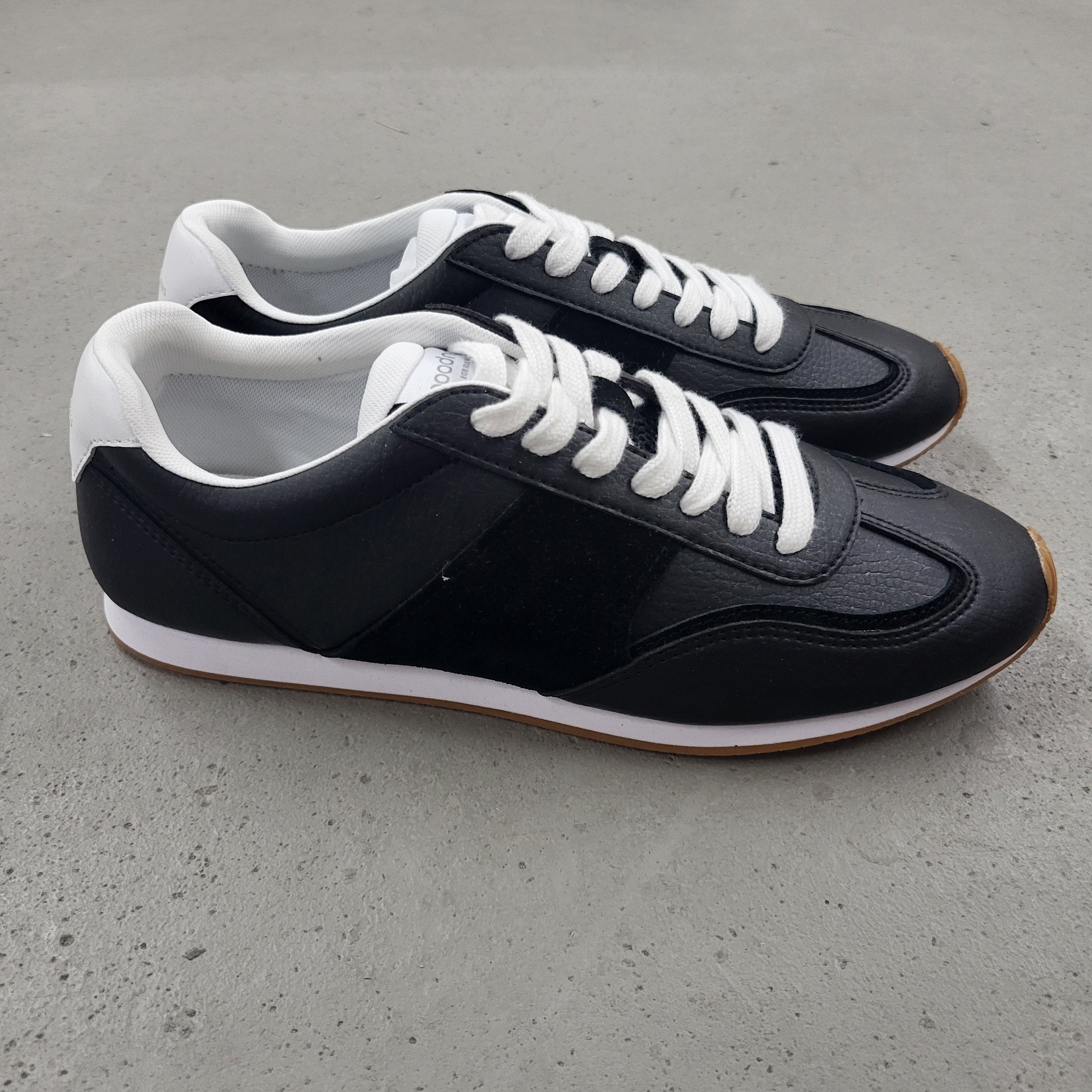 Ein Paar schwarz-weiße Turnschuhe mit weißen Schnürsenkeln steht auf einer glatten grauen Fläche. Die Schuhe haben weiße Sohlen und weiße Akzente an der Ferse und am Kragen.