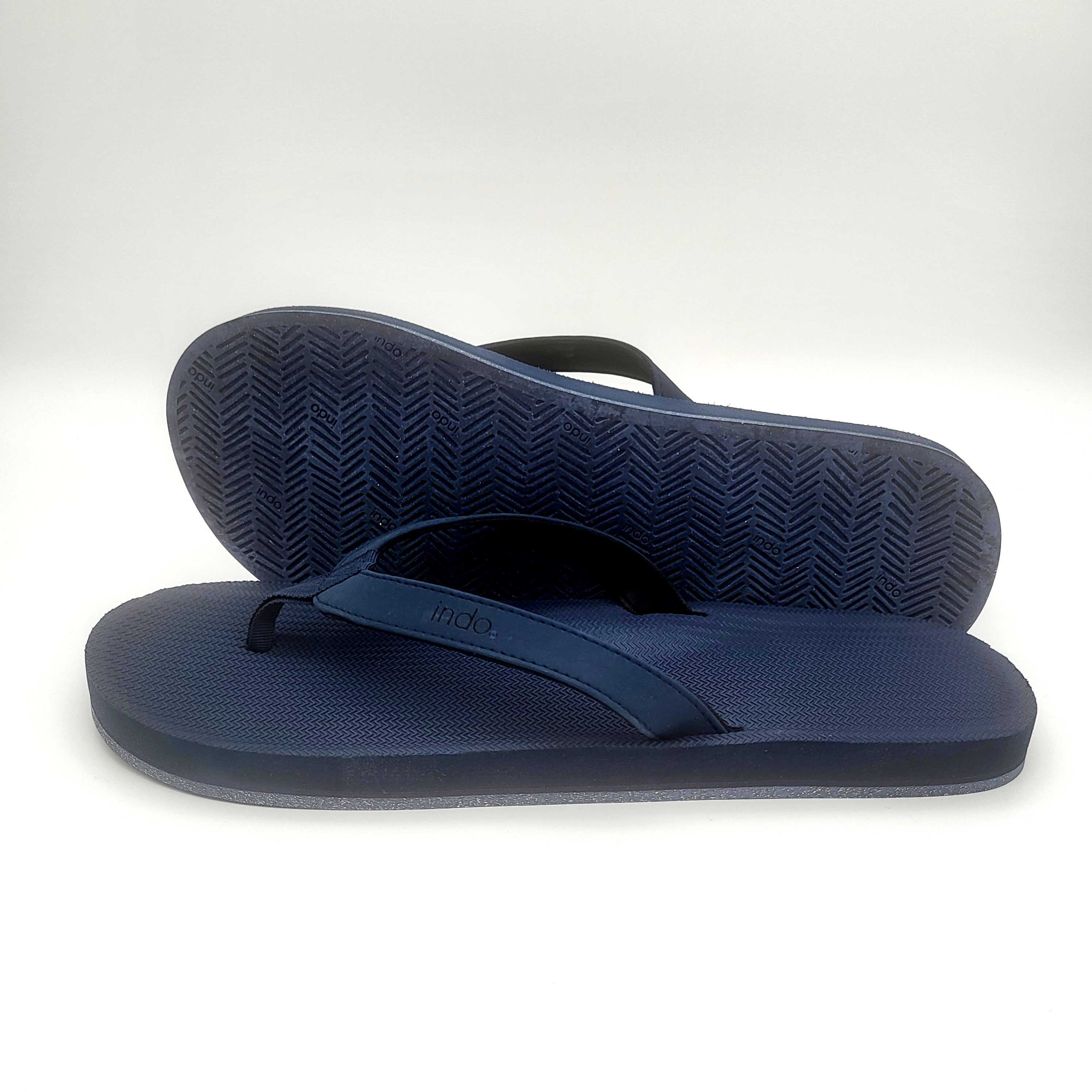 Ein Paar marineblaue Flip-Flops, von denen einer flach liegt und der andere hinter ihm aufgestützt ist, wobei sowohl die strukturierte Sohle als auch das obere Riemenmuster vor einem einfarbig weißen Hintergrund zu sehen sind.