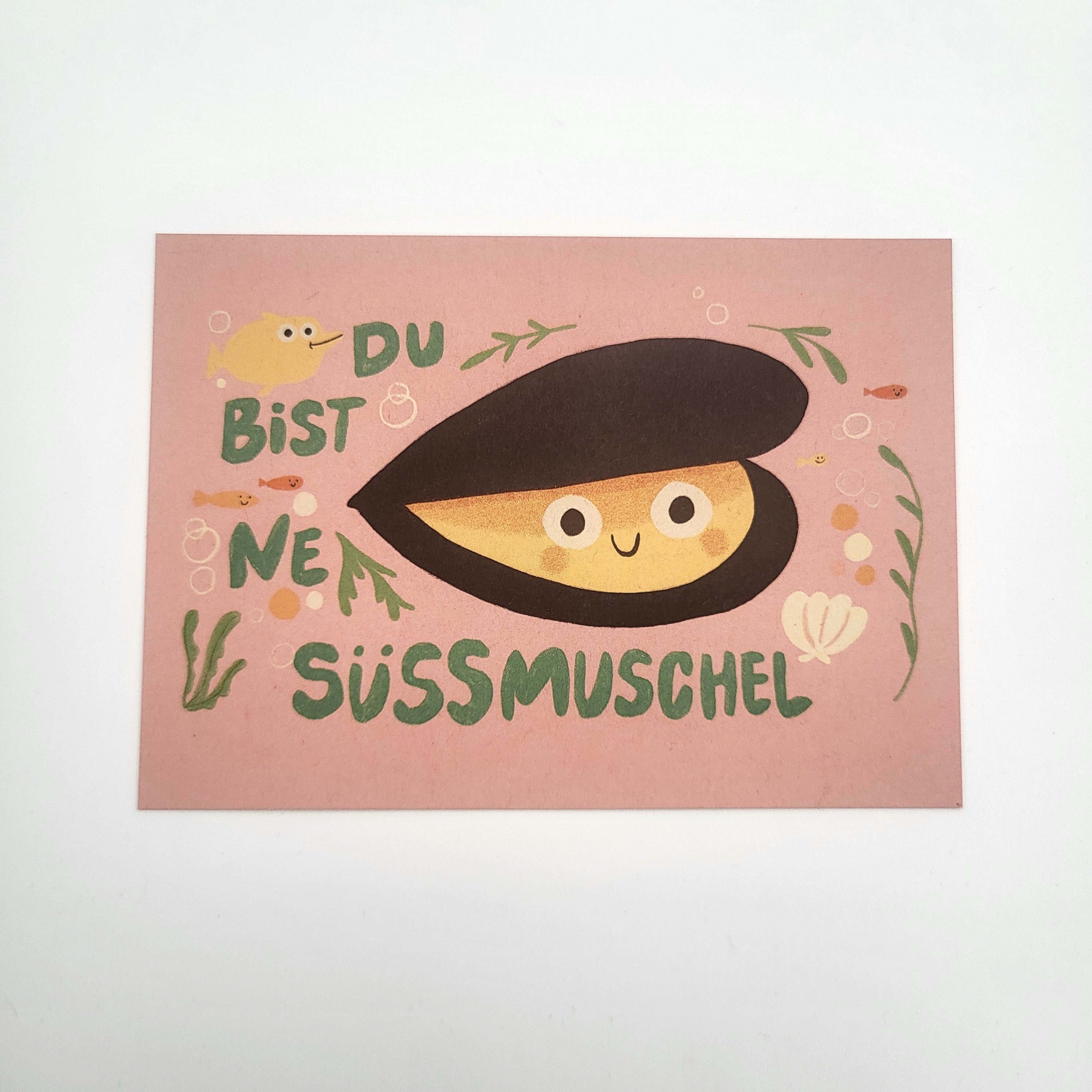 Eine Postkarte mit einer lächelnden Cartoon-Muschel, umgeben von Algen und kleinen Fischen. Der Text auf Deutsch lautet "Du bist ne Süßmuschel" auf einem rosa Hintergrund.