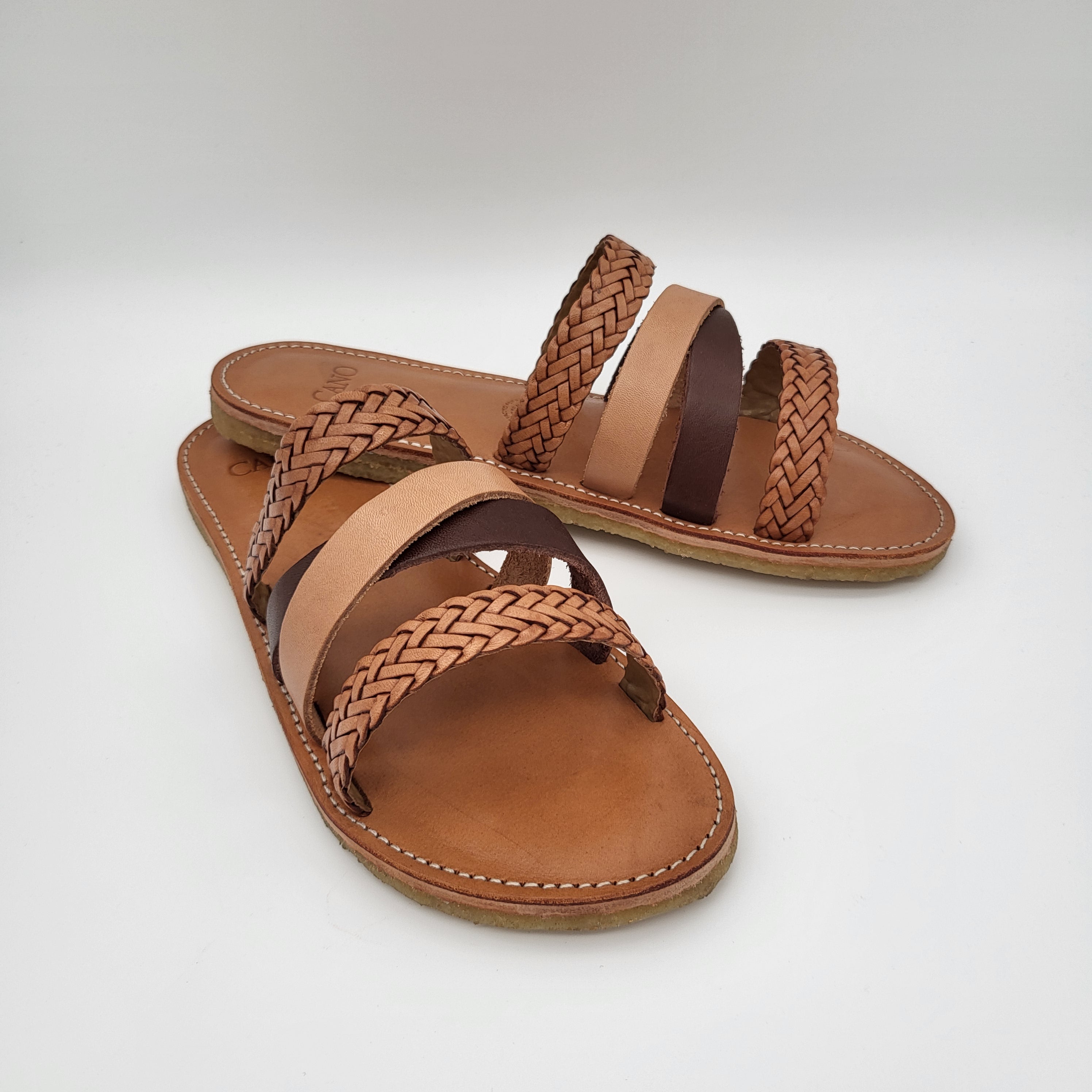 Ein Paar hellbraune Ledersandalen mit geflochtenen und glatten Riemen, die vor einem schlichten weißen Hintergrund präsentiert werden. Die Sandalen haben flache Sohlen und sind zehenoffen.