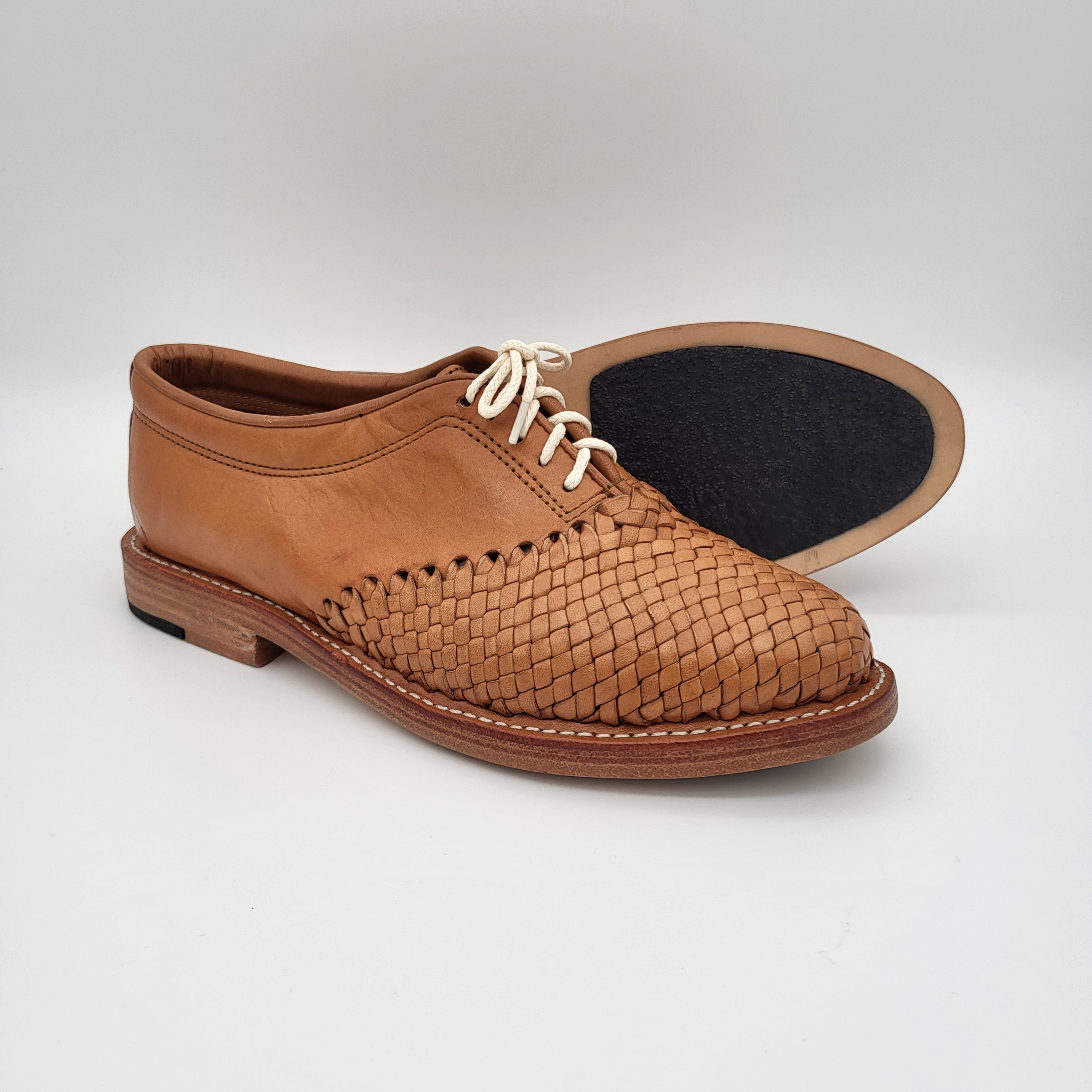 Ein Paar hellbraune Lederschnürschuhe mit einem gewebten Muster auf der Vorderseite und Glattleder an den Seiten, wobei ein Schuh eine schwarze Sohle aufweist.