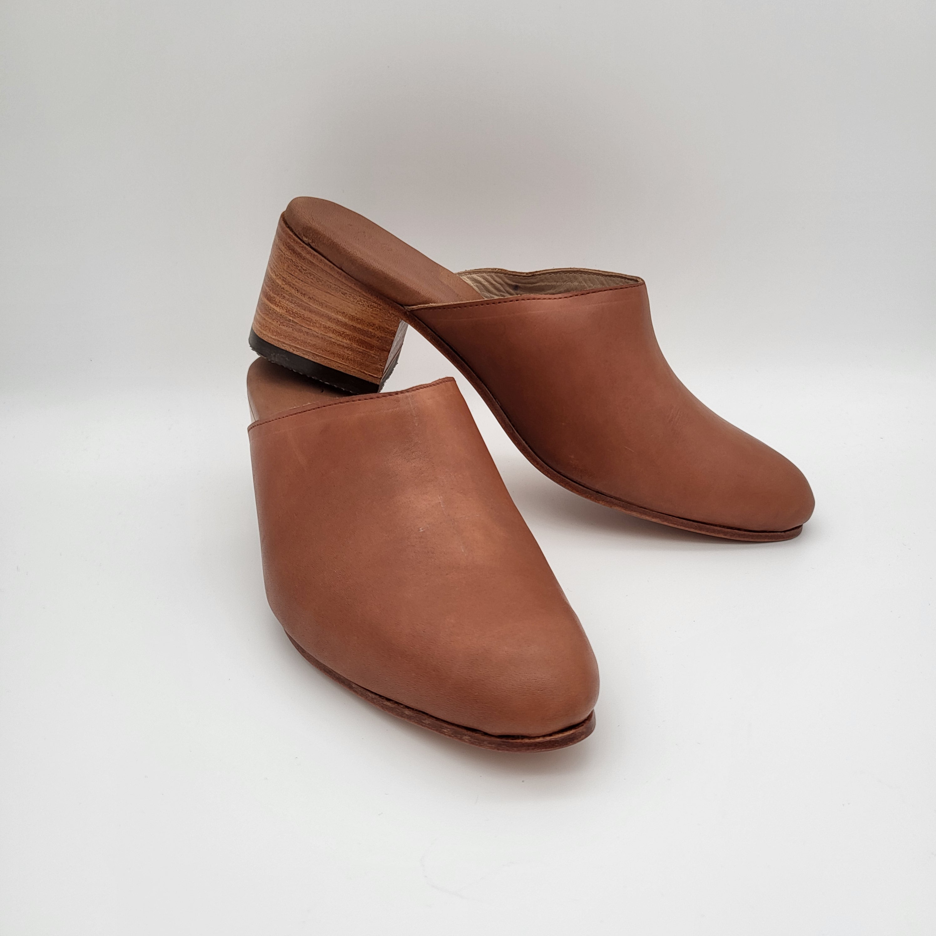 Ein Paar braune Lederpantoletten mit geschlossenen Zehen und klobigen Holzabsätzen auf einem weißen Hintergrund.