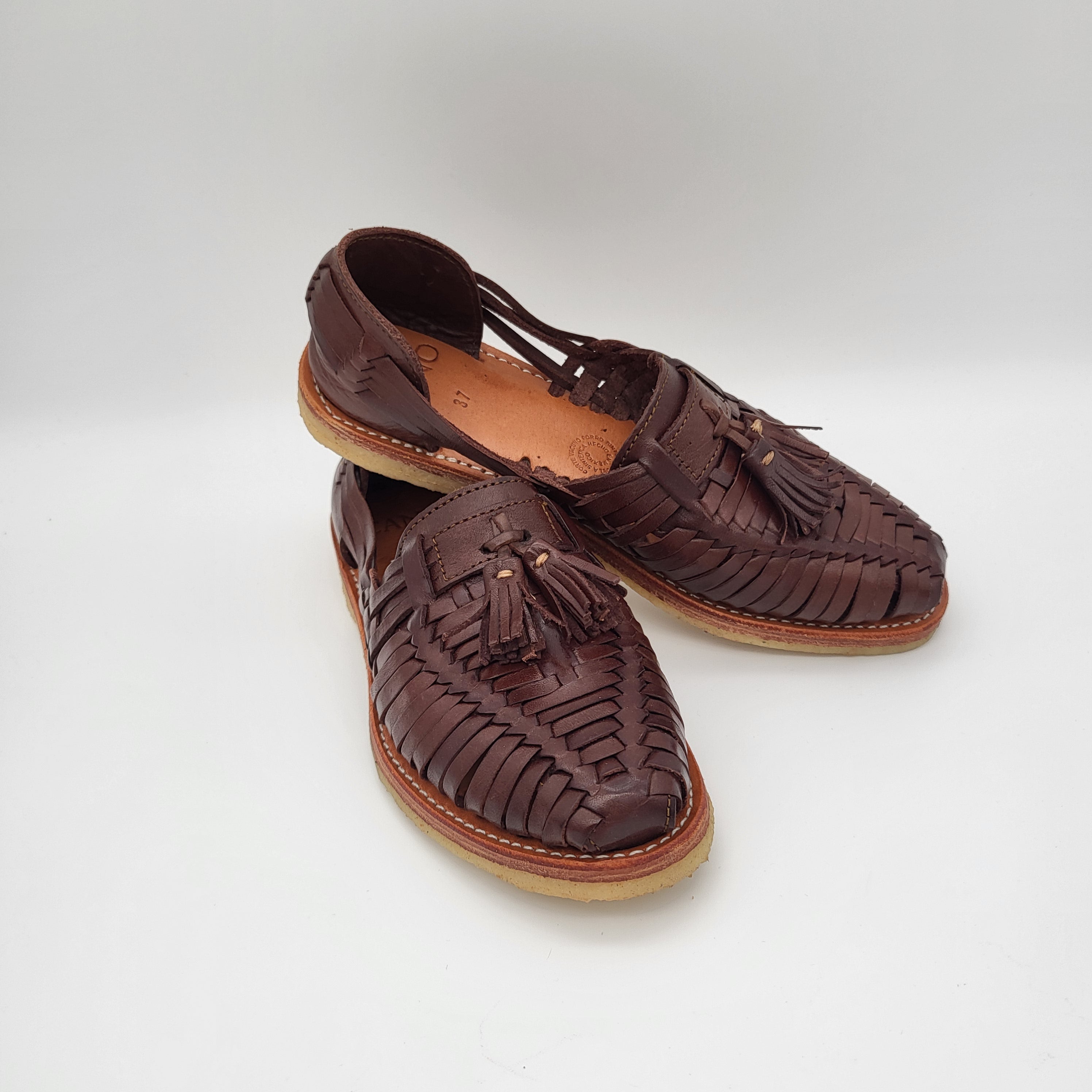 Ein Paar braune Sandalen aus gewebtem Leder mit Quasten auf der Vorderseite, die vor einem schlichten weißen Hintergrund stehen.