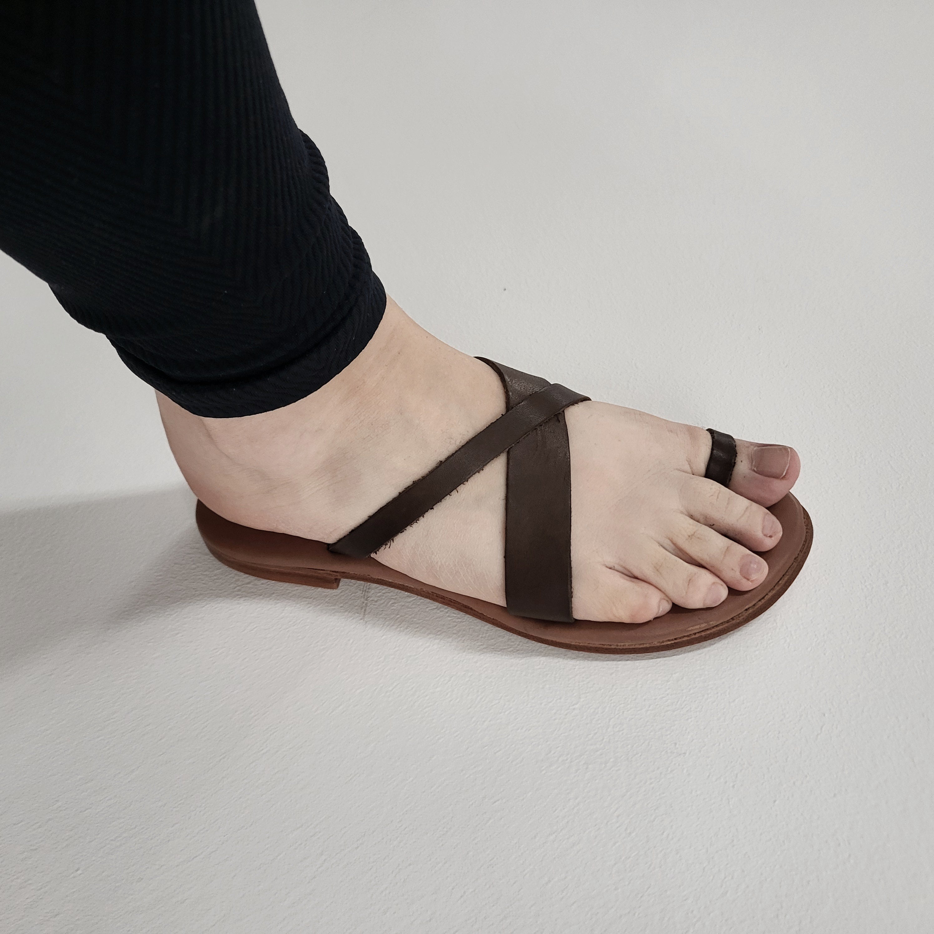 Eine Person, die eine schwarze Hose und eine braune Sandale mit gekreuzten Riemen und einer Schlaufe um den großen Zeh trägt, steht auf einem hellen Boden.