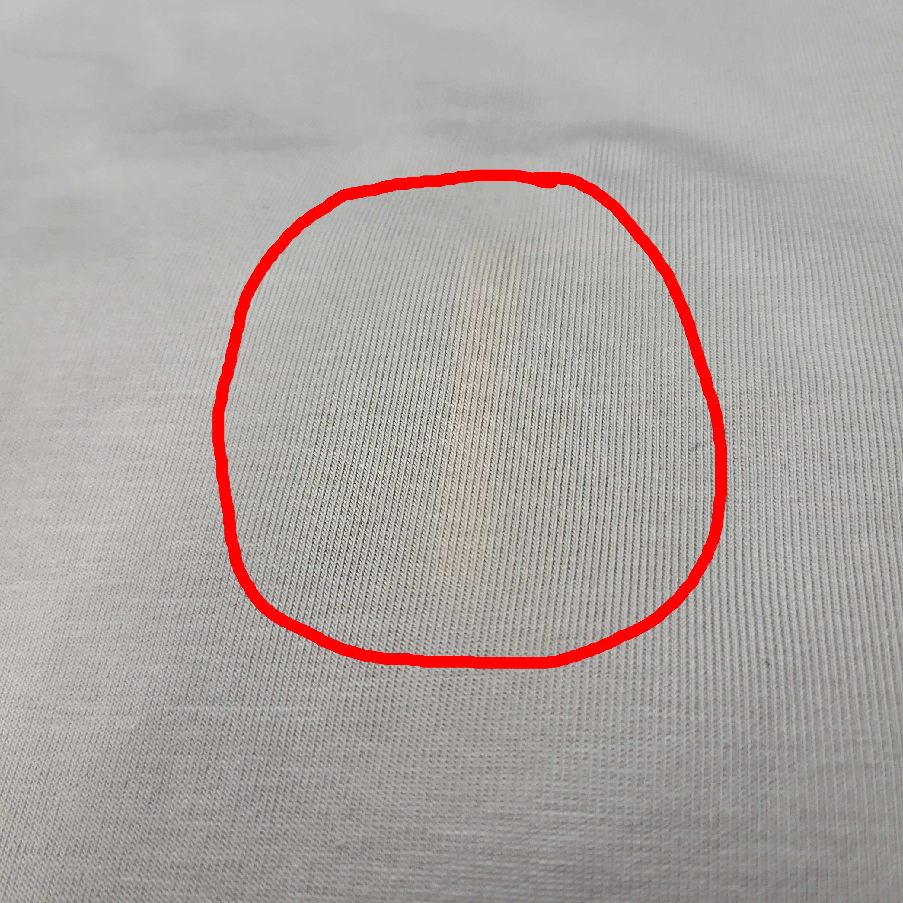 Nahaufnahme eines weißen Gewebes mit einem schwachen, schmalen, gelblichen Fleck, der nahe der Mitte vertikal verläuft. Die Gewebestruktur und die Bindung sind sichtbar.