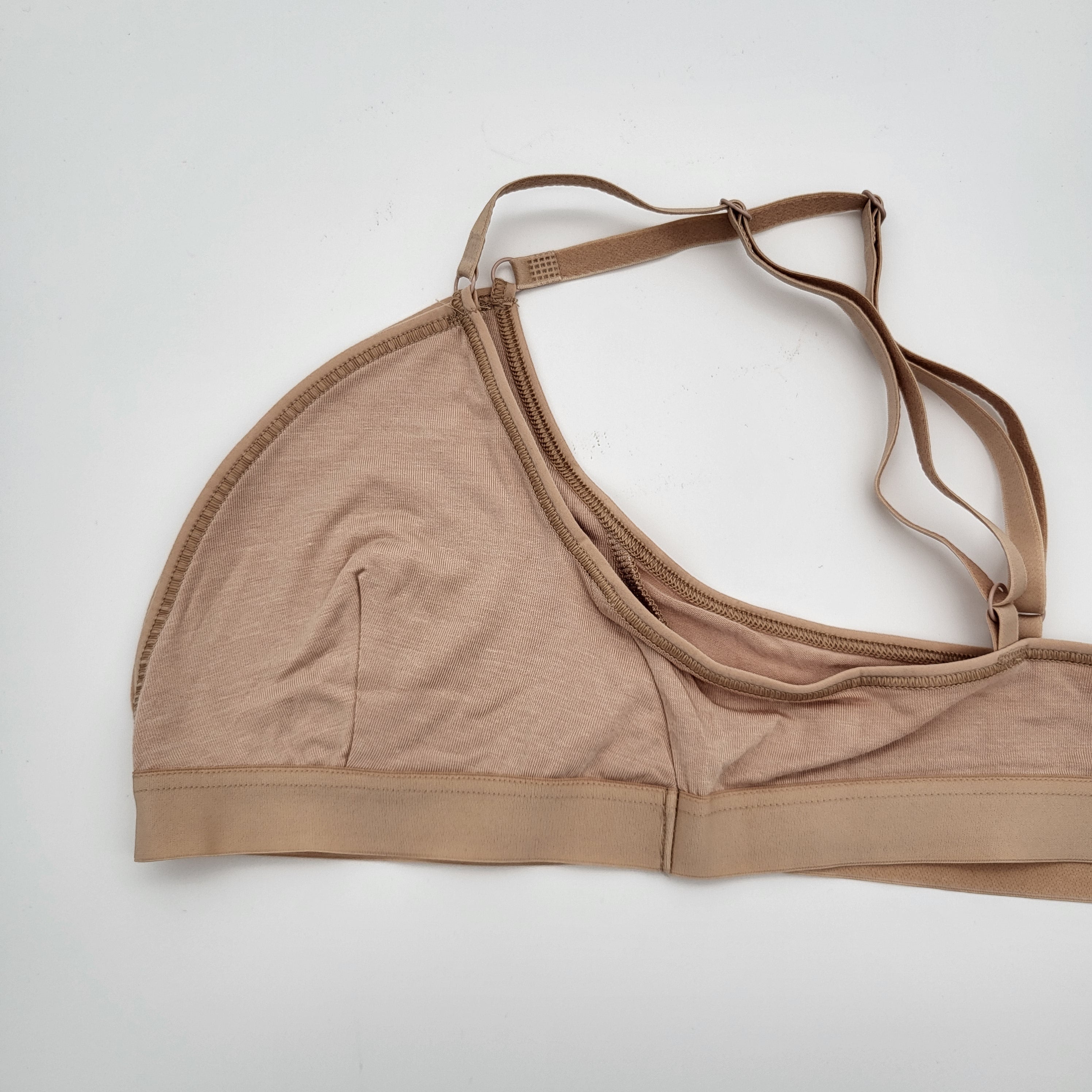 Ein hellbrauner oder beiger Bralette mit verstellbaren Trägern liegt flach auf einer weißen Fläche. Der Bralette ist ungefüttert, aus weichem Stoff und hat ein einfaches, minimalistisches Design.