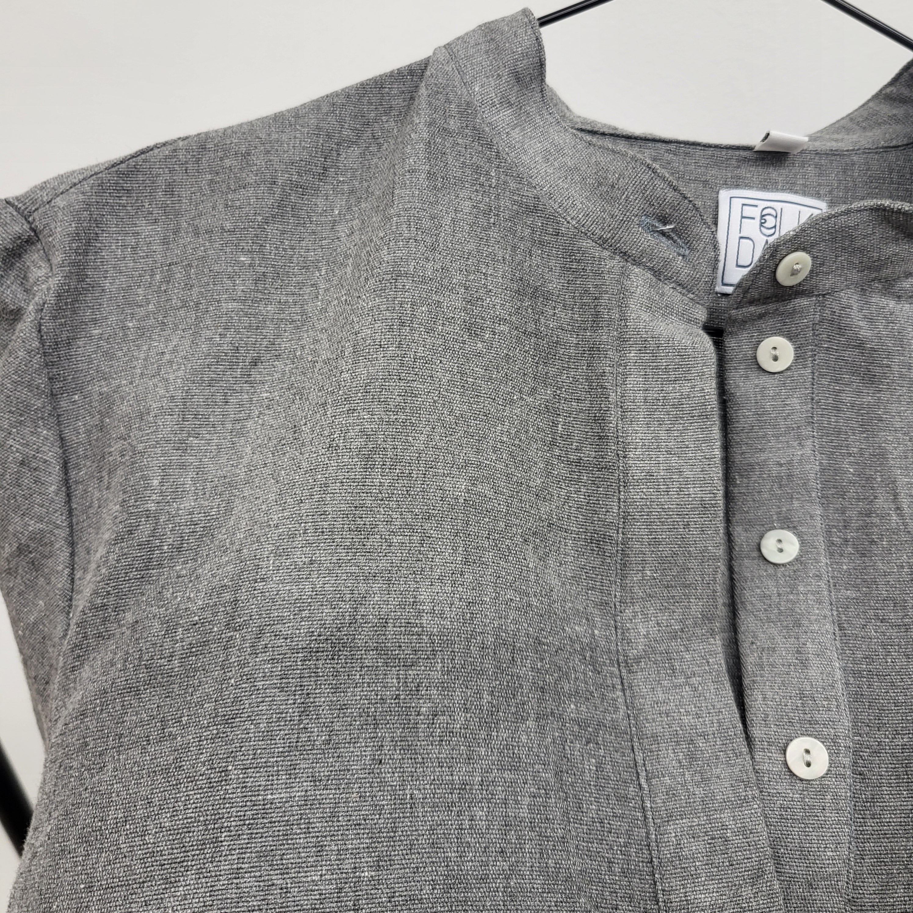 Nahaufnahme eines grauen, strukturierten Hemdes mit vier weißen Knöpfen und einem Stehkragen, das auf einem Bügel vor einem schlichten Hintergrund hängt.