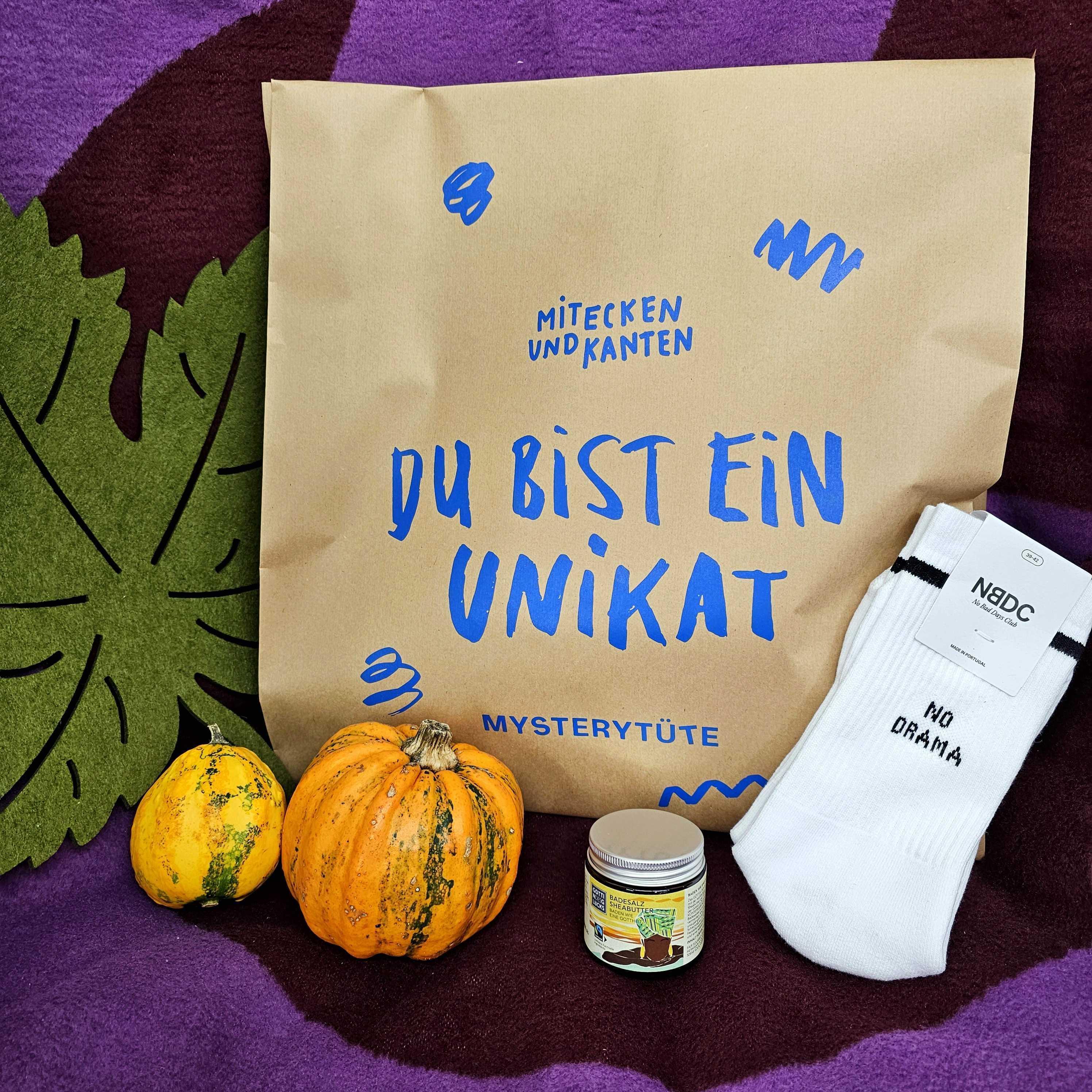 Eine braune Papiertüte mit blauer Schrift, zwei kleine Kürbisse, ein Paar weiße Socken mit der Aufschrift NO DRAMA und ein kleines Glas sind auf einem lila und grünen Hintergrund angeordnet.