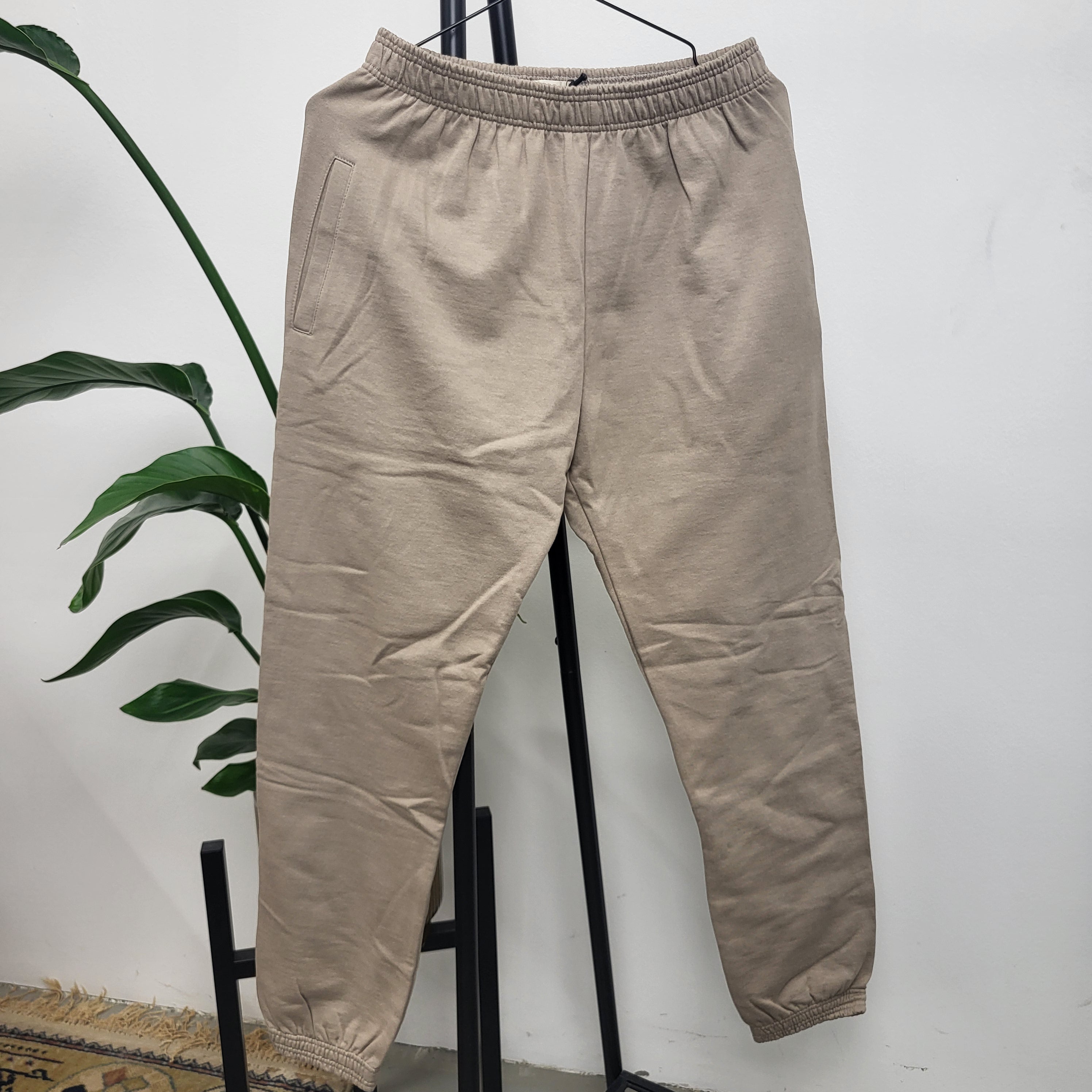 Eine beigefarbene Jogginghose mit elastischem Bund und Bündchen hängt an einem schwarzen Ständer. Eine grüne Blattpflanze und ein Teil eines gemusterten Teppichs sind im Hintergrund vor einer weißen Wand zu sehen.