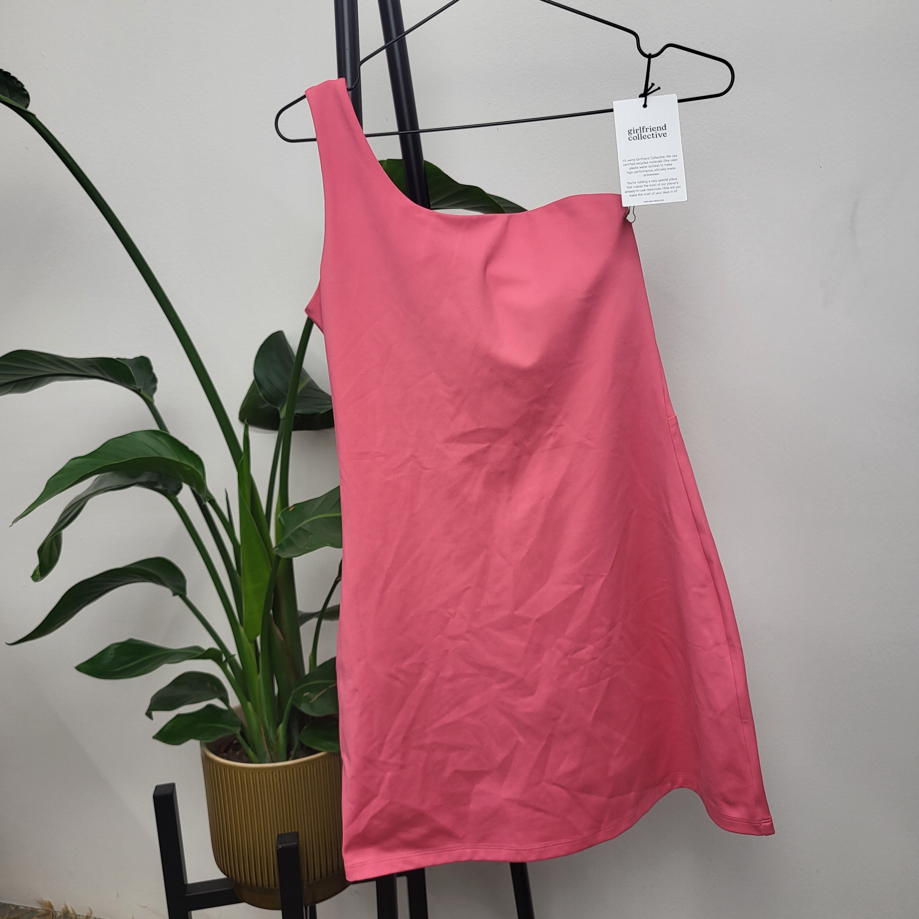 Ein hellrosa, einschulteriges Kleid hängt auf einem schwarzen Kleiderbügel vor einer weißen Wand. Daneben steht eine grüne Topfpflanze, und an dem Kleid ist ein weißes Schild mit Text befestigt.