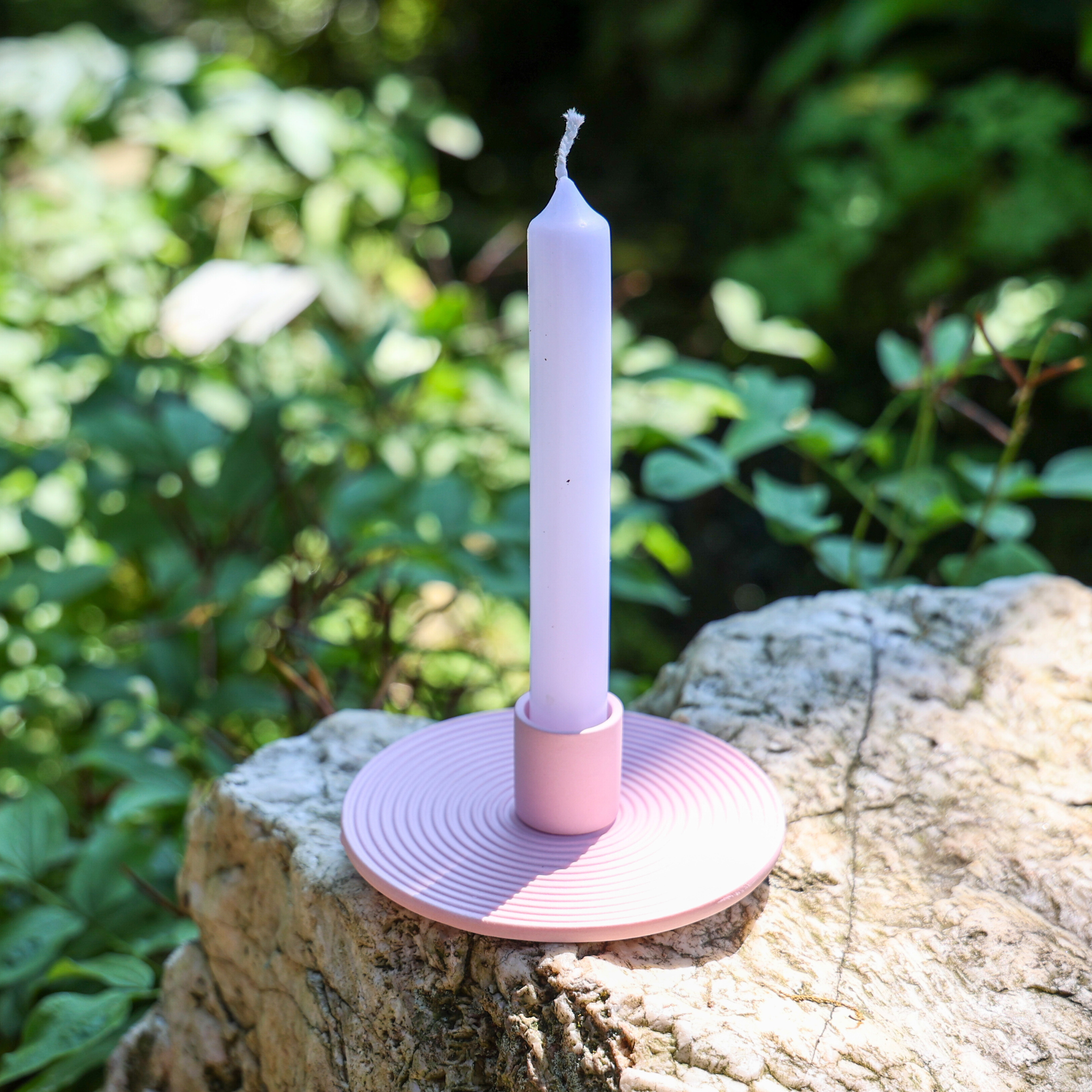 Eine lavendelfarbene Kegelkerze in einem hellrosa Halter sitzt auf einem strukturierten Felsen im Freien, umgeben von grünen Pflanzen im Sonnenlicht.