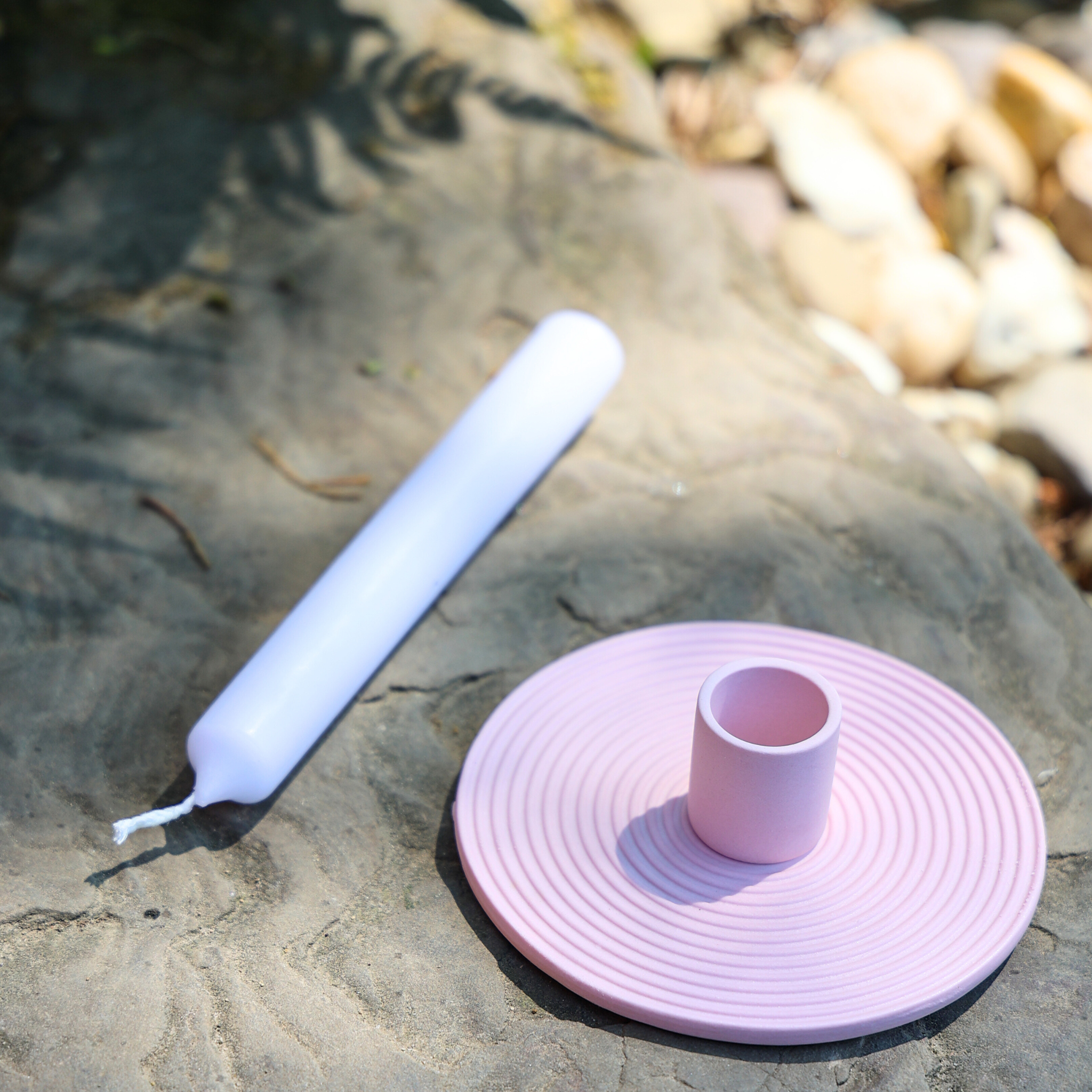 Eine weiße Spitzkerze und ein rosafarbener Keramikkerzenhalter stehen auf einer Steinfläche im Freien, wobei das Sonnenlicht Schatten wirft und im Hintergrund Kieselsteine zu sehen sind.