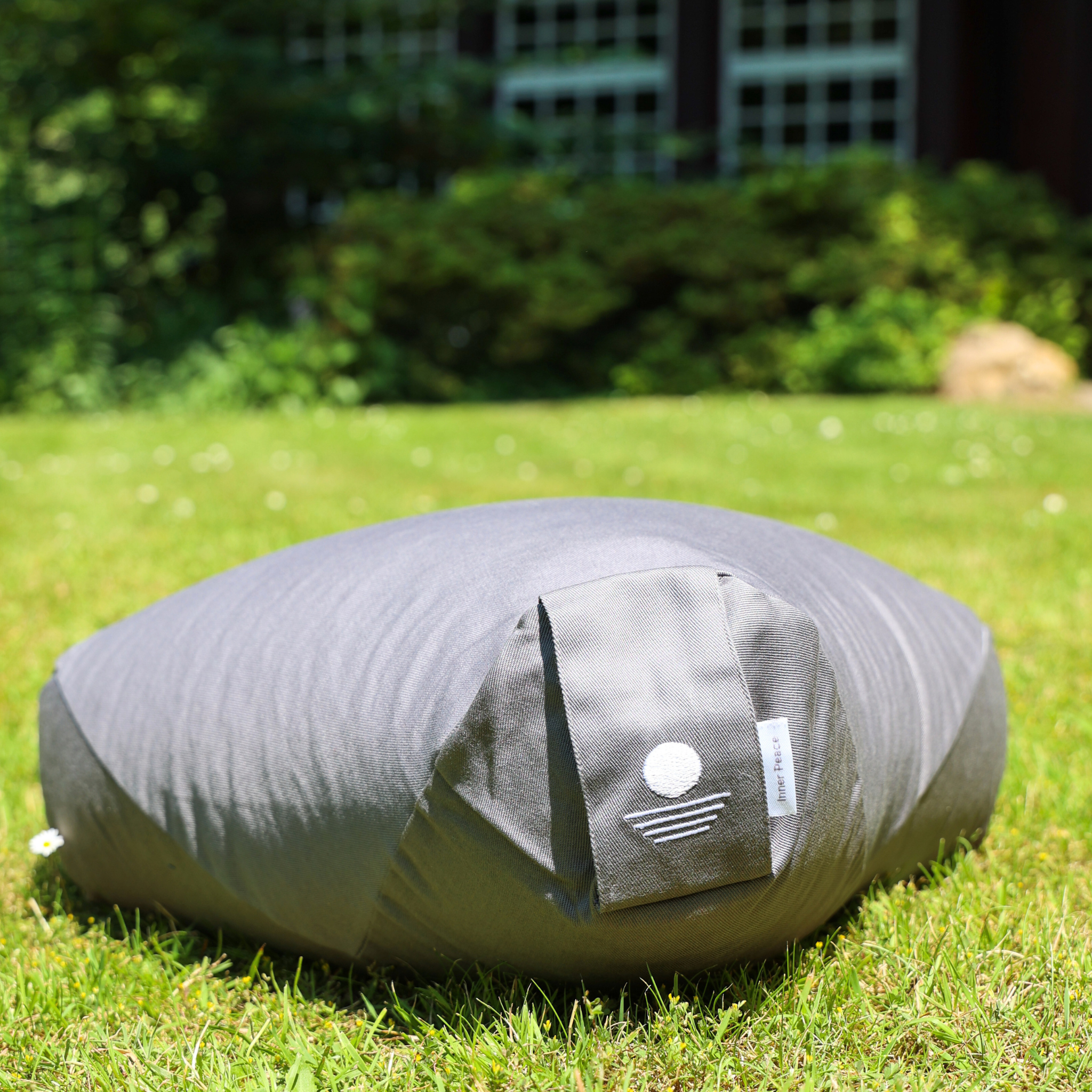 Ein grauer Outdoor-Sitzsack ruht auf grünem Gras in einem sonnigen Garten, mit Bäumen und einem Gebäude im unscharfen Hintergrund. Ein weißes Logo ist auf der Tasche des Sitzsacks zu sehen.