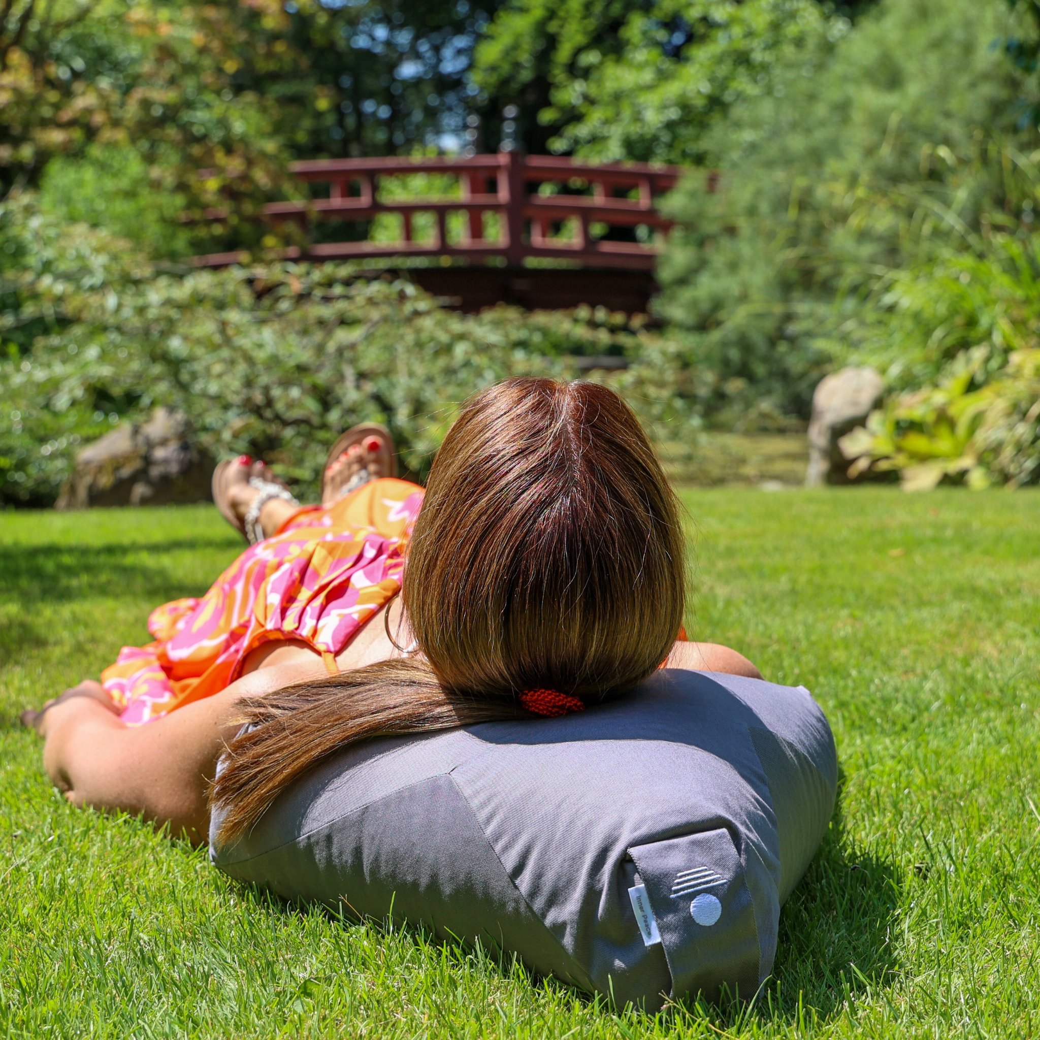 Eine Person mit hellbraunem Haar liegt auf einem grauen Kissen auf grünem Gras, trägt ein buntes Kleid und blickt von der Kamera weg auf eine Holzbrücke und einen üppigen Garten.