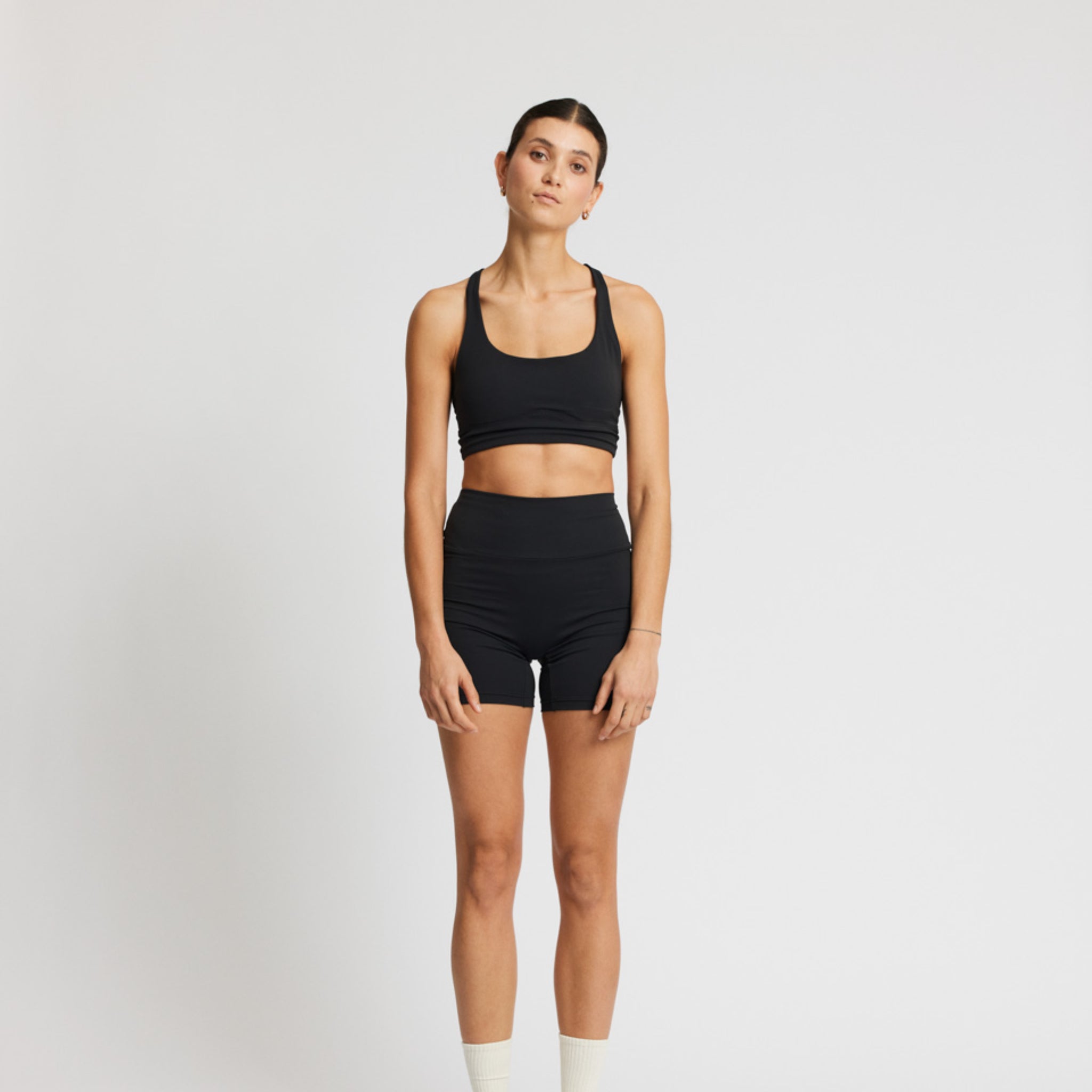 Frau in schwarzer Sportbekleidung mit Shorts und Crop-Top steht vor einem weißen Hintergrund, frontal zur Kamera.