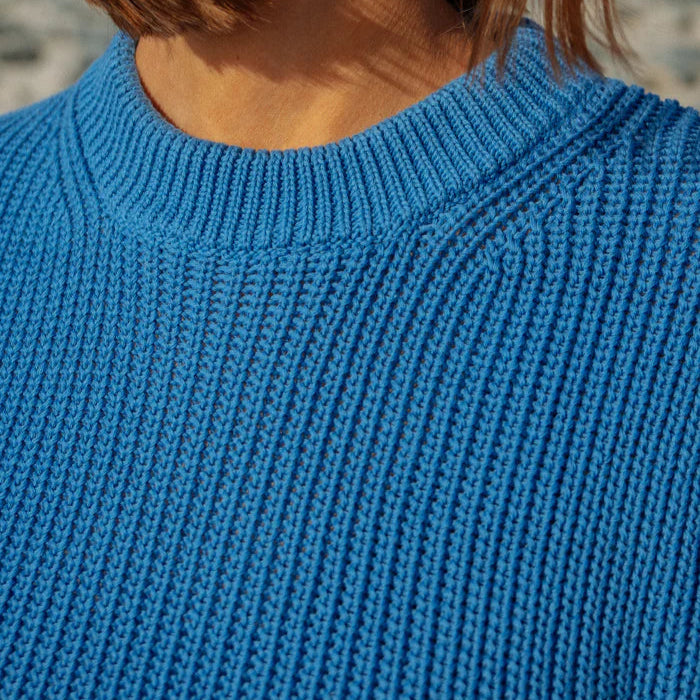 Nahaufnahme einer Person, die einen hellblauen, gerippten Strickpullover trägt. Nur der untere Teil des Gesichts, der Hals und die Haare sind zu sehen; der Hintergrund ist unscharf.