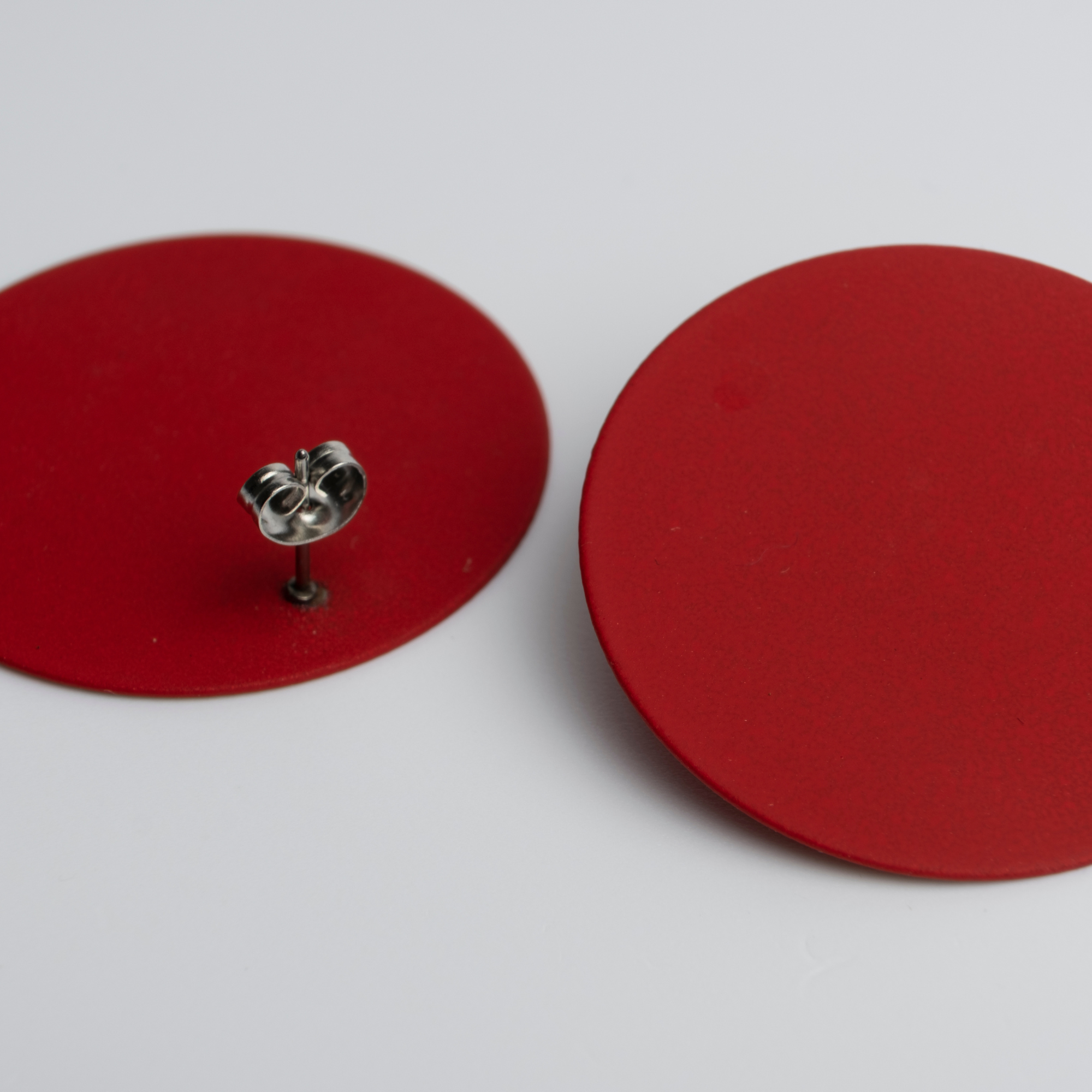 Zwei große, flache, runde rote Ohrringe sind auf einer weißen Fläche ausgestellt. Ein Ohrring zeigt den silbernen Stift und die Halterung, während der andere mit der Vorderseite nach oben liegt.
