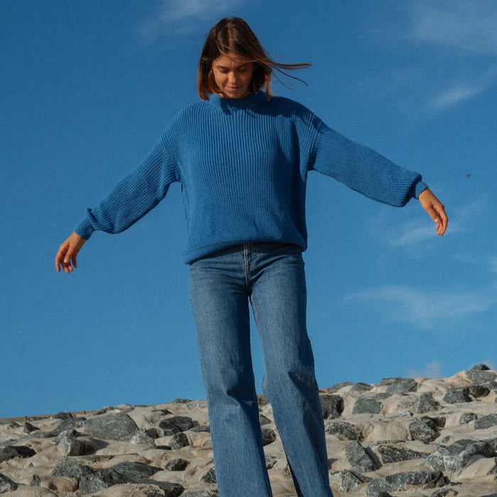 Eine Frau, die einen blauen Pullover und Jeans trägt, steht mit leicht ausgestreckten Armen auf einer felsigen Fläche unter einem klaren blauen Himmel.