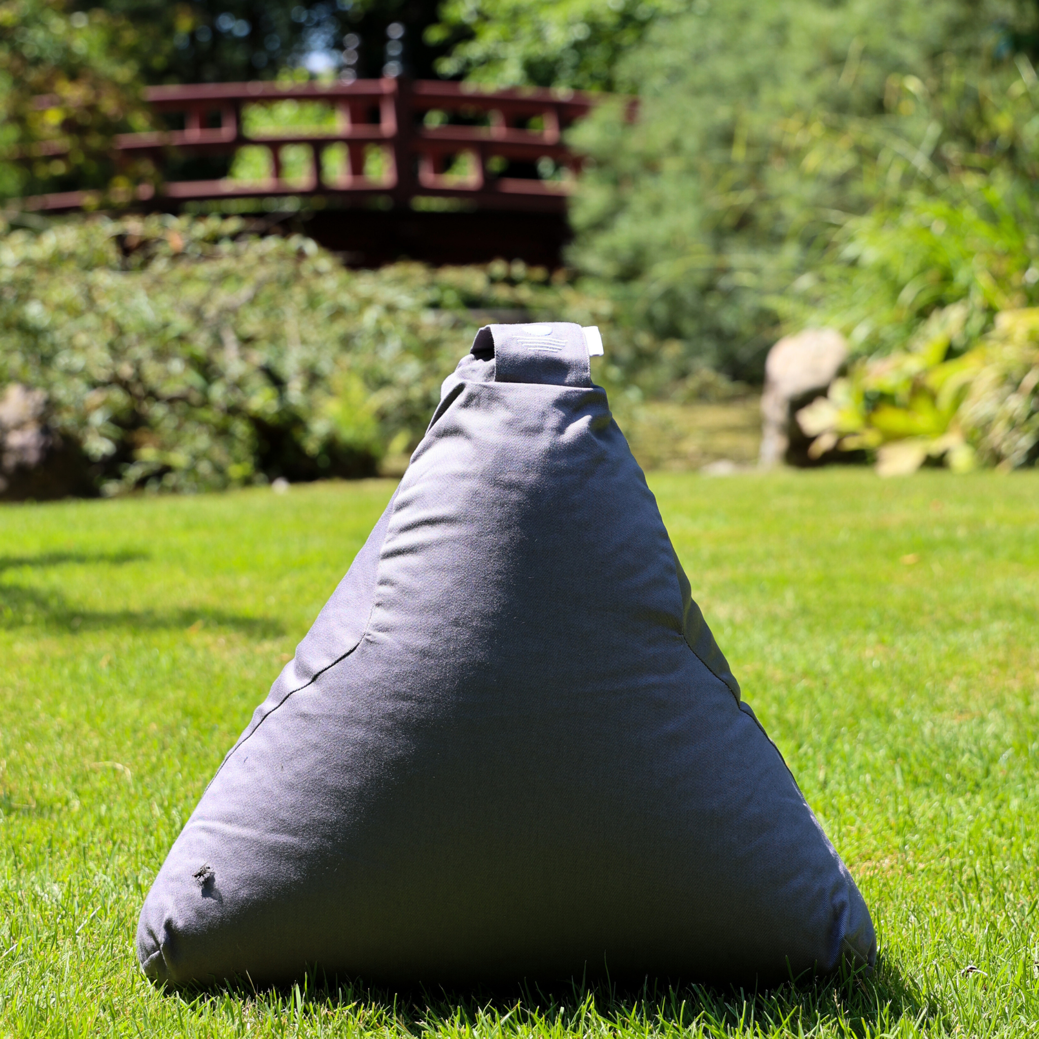 Ein grauer, dreieckiger Sitzsack sitzt auf grünem Gras in einem sonnenbeschienenen Garten, in dessen Hintergrund Bäume und eine rote Holzbrücke verschwimmen.