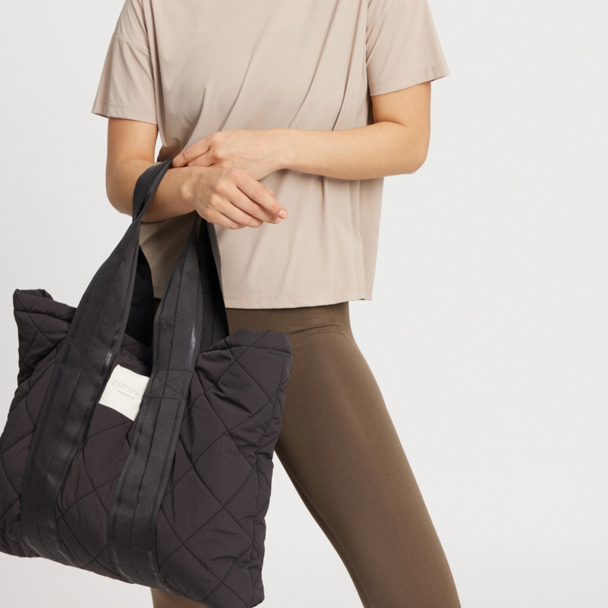 Person in beige T-Shirt und brauner Leggings hält eine große, schwarze gepolsterte Tasche mit Griffen.