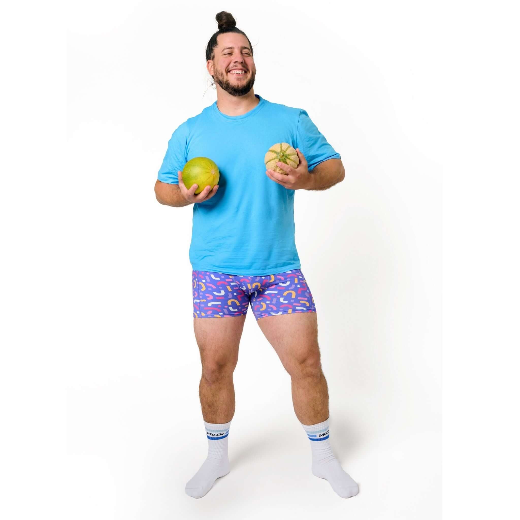 Mann in blauem T-Shirt und gemusterten Shorts hält zwei Melonen und lächelt vor weißem Hintergrund.