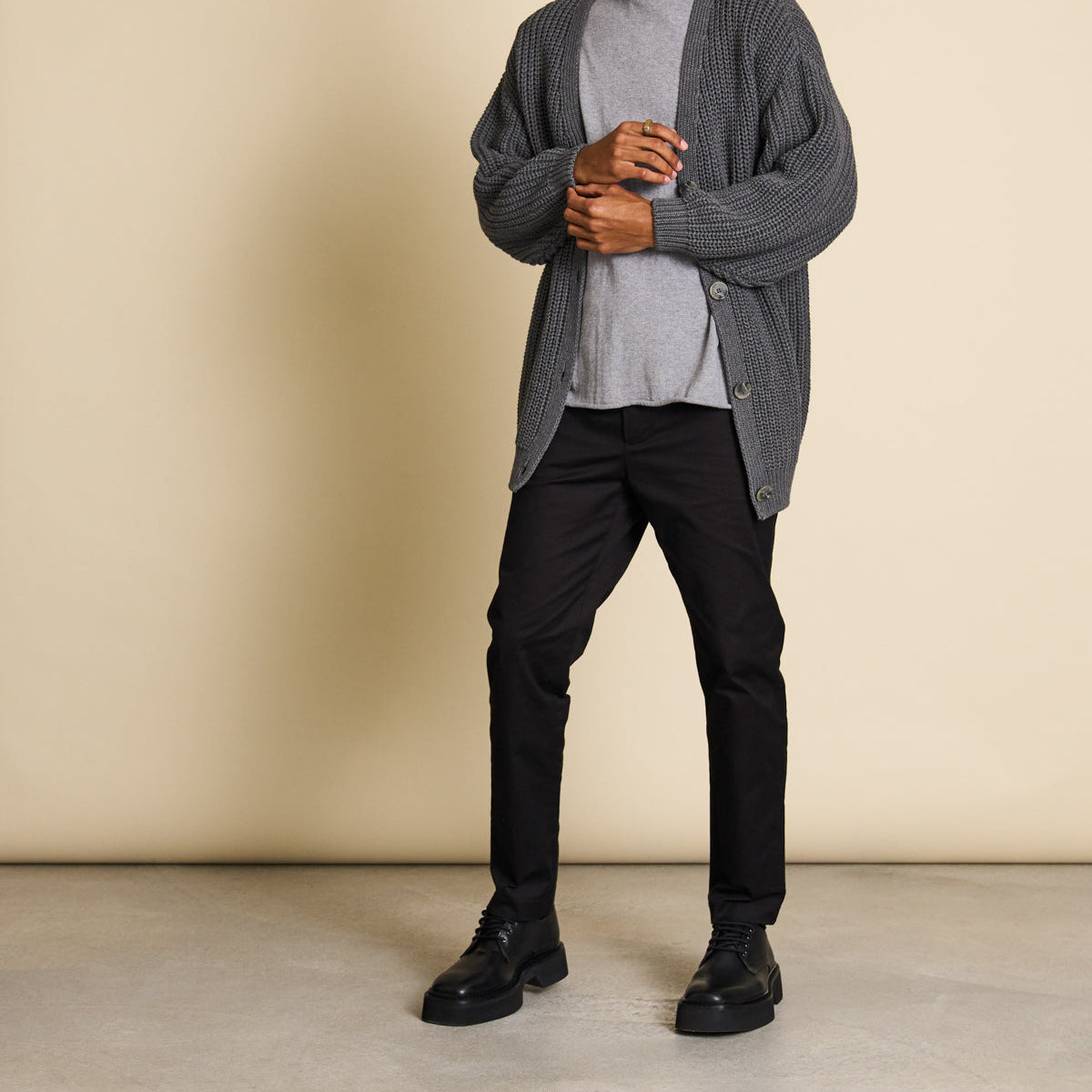 Person mit grauer Strickjacke, grauem Shirt, schwarzen Hosen und schwarzen Schuhen vor beigem Hintergrund.