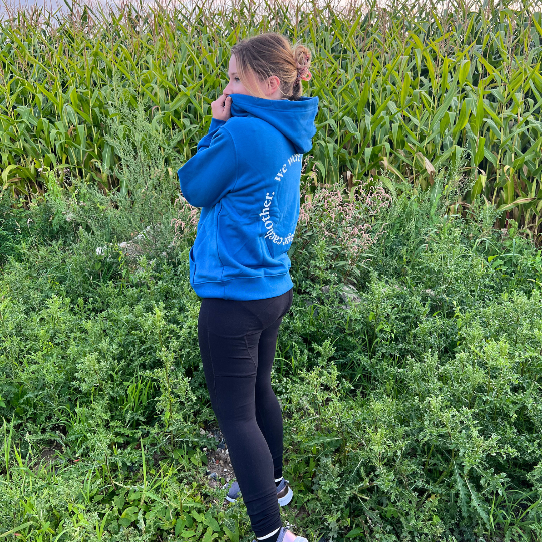 Eine Person im blauen Hoodie steht in einer grünen Landschaft vor einem Maisfeld.