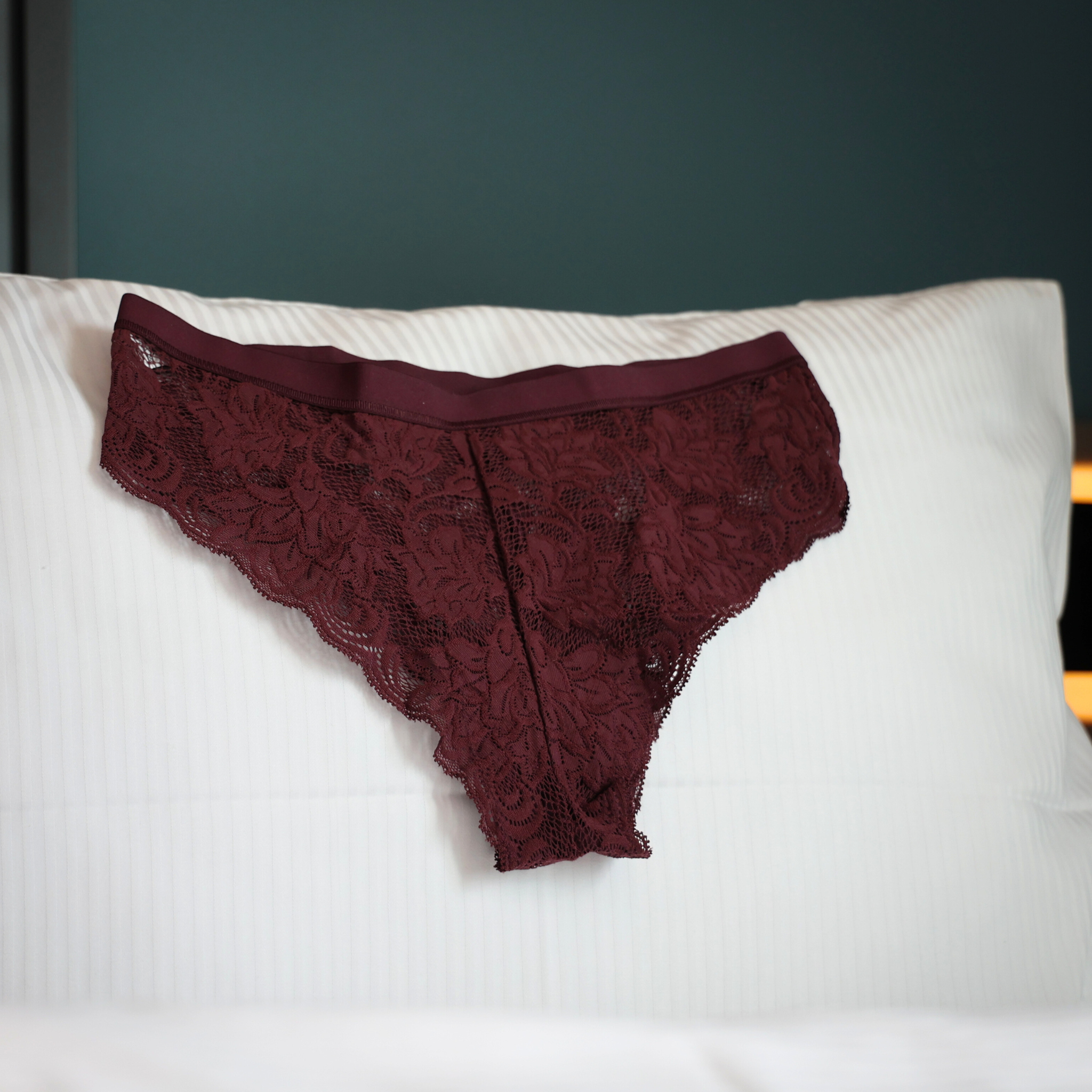 Ein weinrotes Spitzenhöschen liegt fein säuberlich auf einem weißen, gestreiften Kissen auf einem Bett, mit einer dunkelgrünen Wand im Hintergrund.